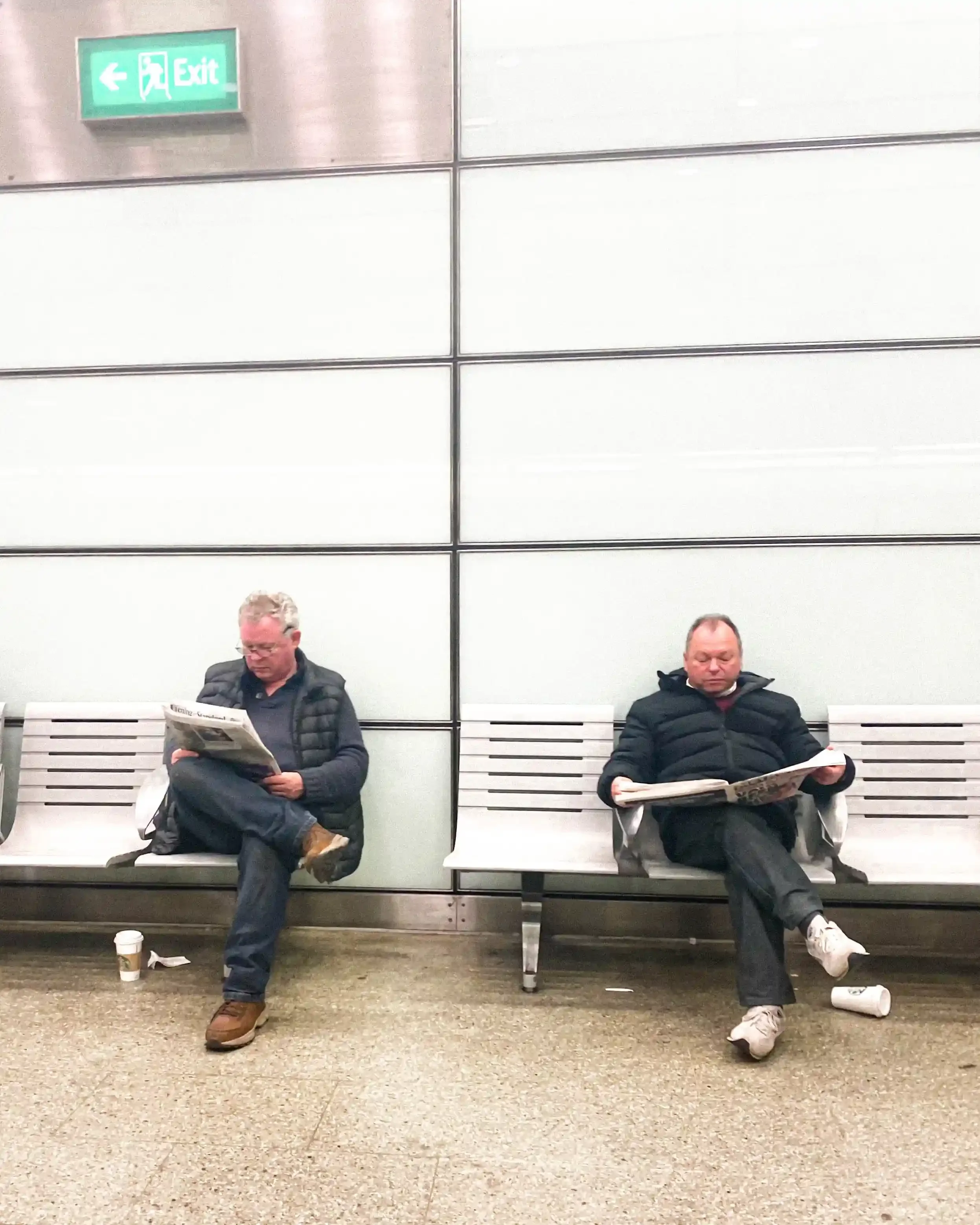 Two men sitting separately on metal station benches, both crossing their legs and reading newspapers under an exit sign, by Matthew Morgan for 'London's Free Newspapers'.