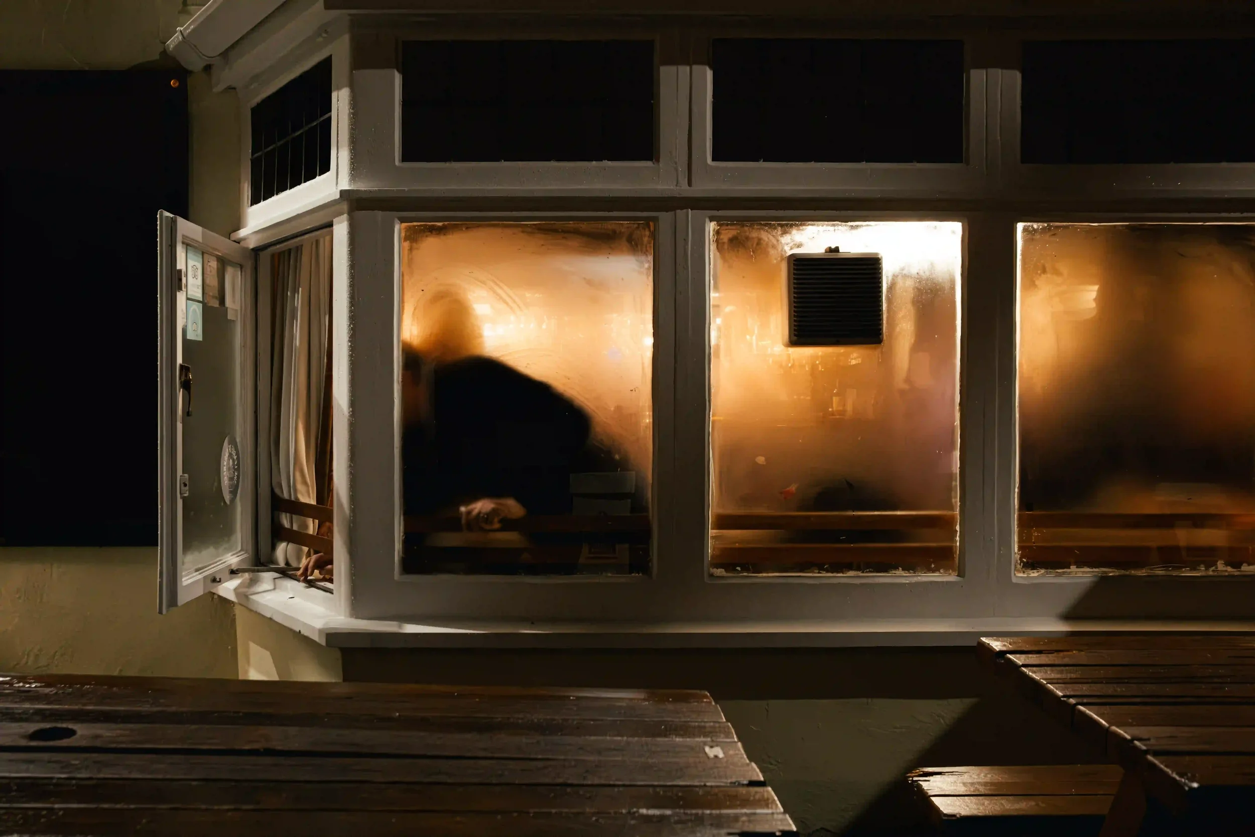 A night view of a white-framed pub bay window with three large panes heavily obscured by golden-orange condensation; a person's hand is visible reaching through a small open side window, with dark wooden picnic tables in the foreground, by Matthew Mo