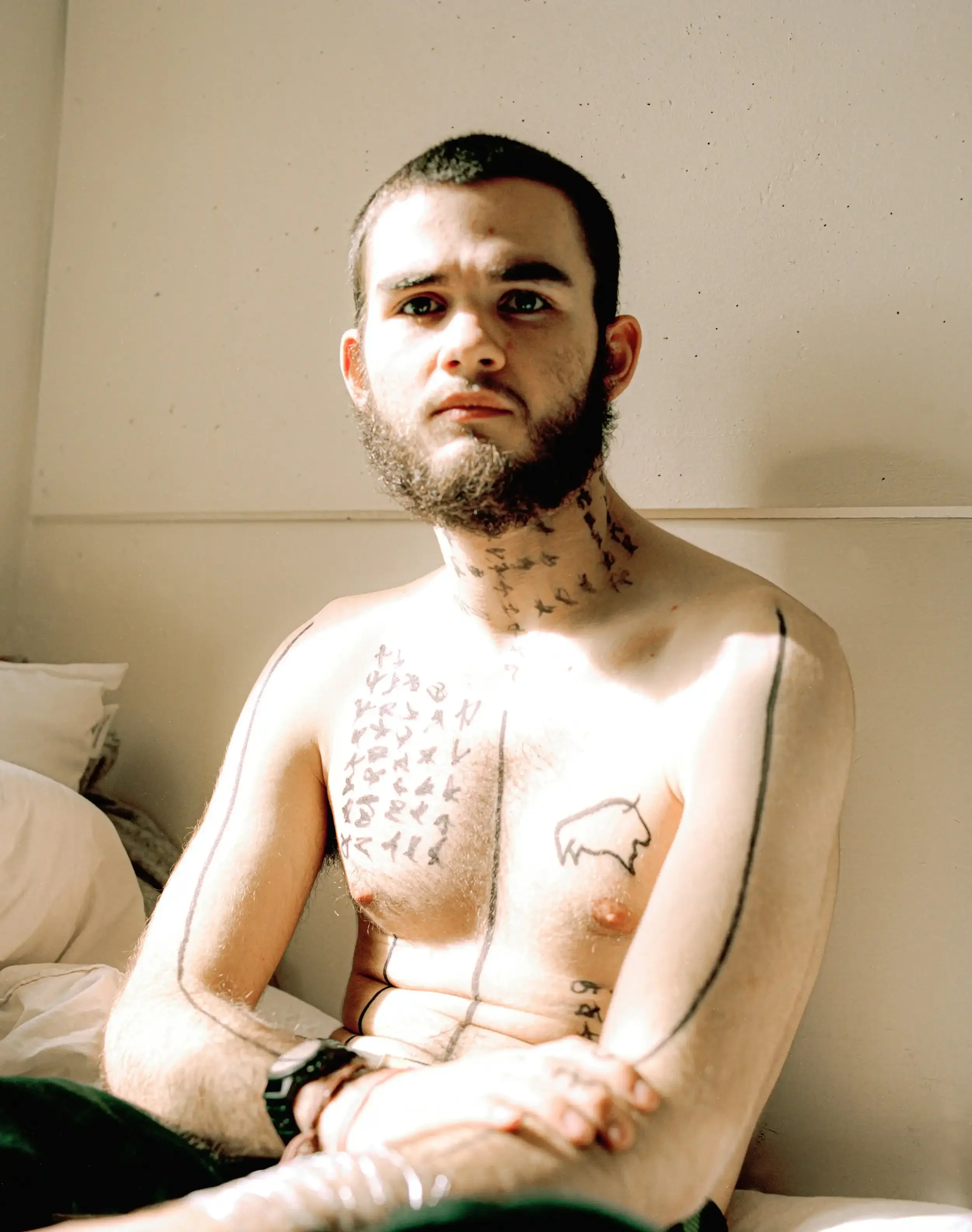 A medium portrait of artist Finlay Cummins, a young man with a short buzz cut and a dark beard, sitting shirtless in a sunlit room. His neck, chest, and arms are covered in intricate, hand-drawn black symbols and bold vertical lines. He looks directl