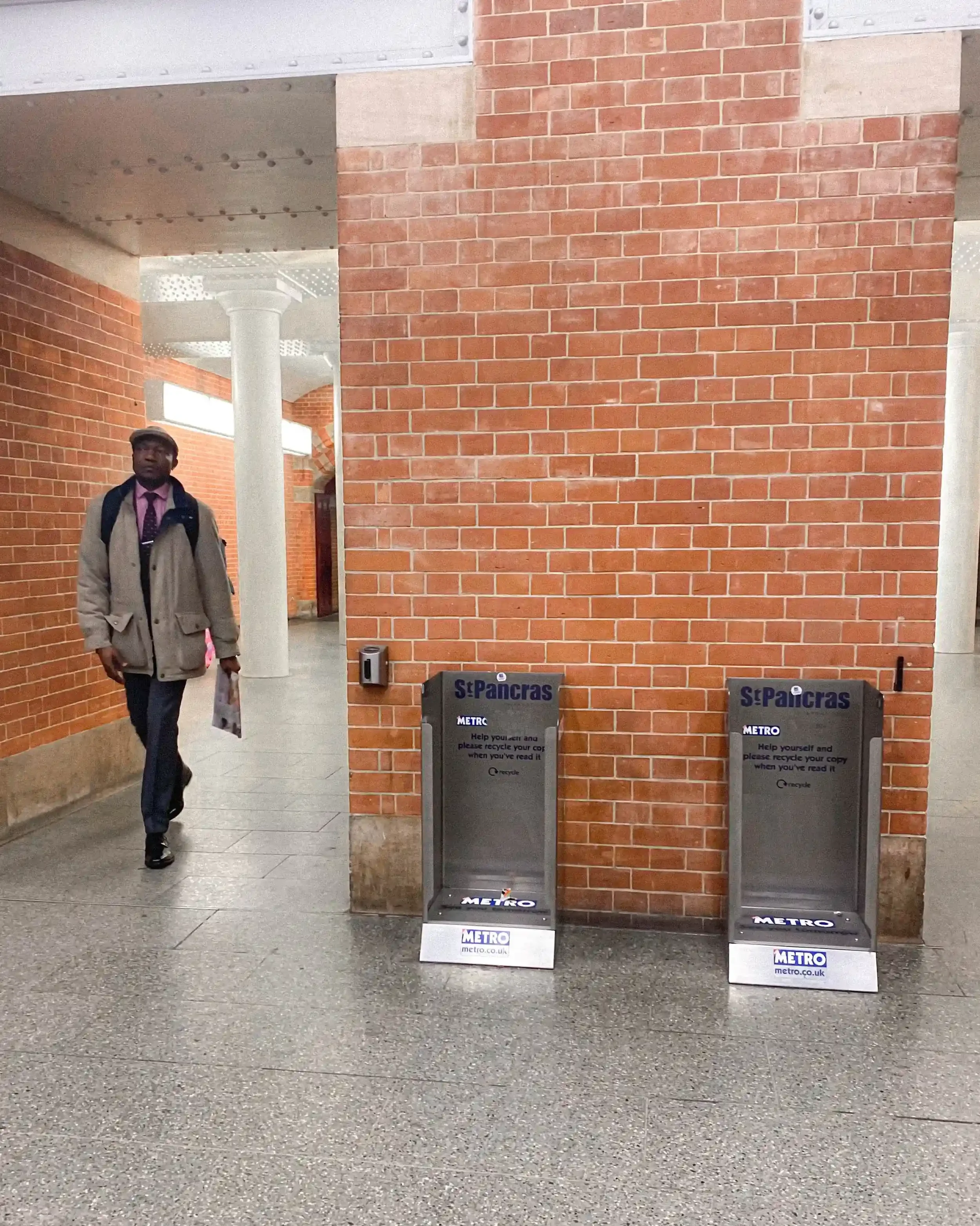 A man in a tan coat walks past empty silver Metro newspaper bins at St Pancras station, holding a newspaper, part of Matthew Morgan's 2022 London documentary series.