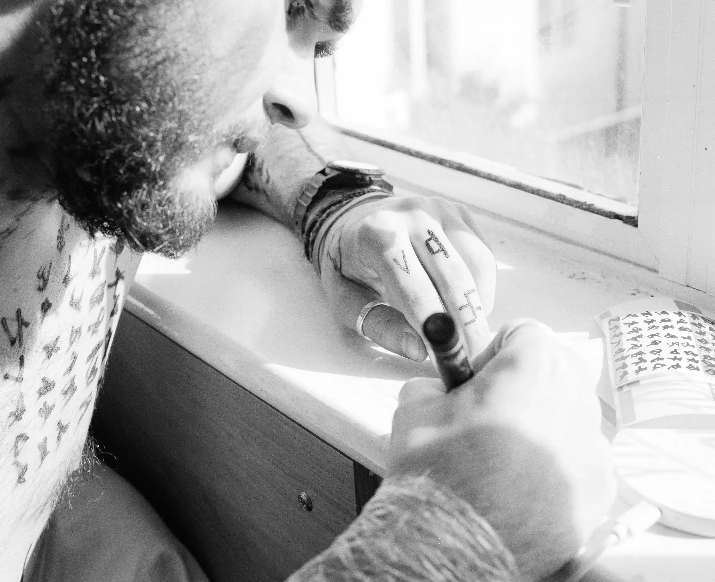 A high-contrast black-and-white close-up of artist Finlay Cummins leaning over a desk by a window. He is using a thick marker to draw small, cryptic symbols onto a sheet of stickers. His hands and chest are covered in similar hand-drawn markings, and