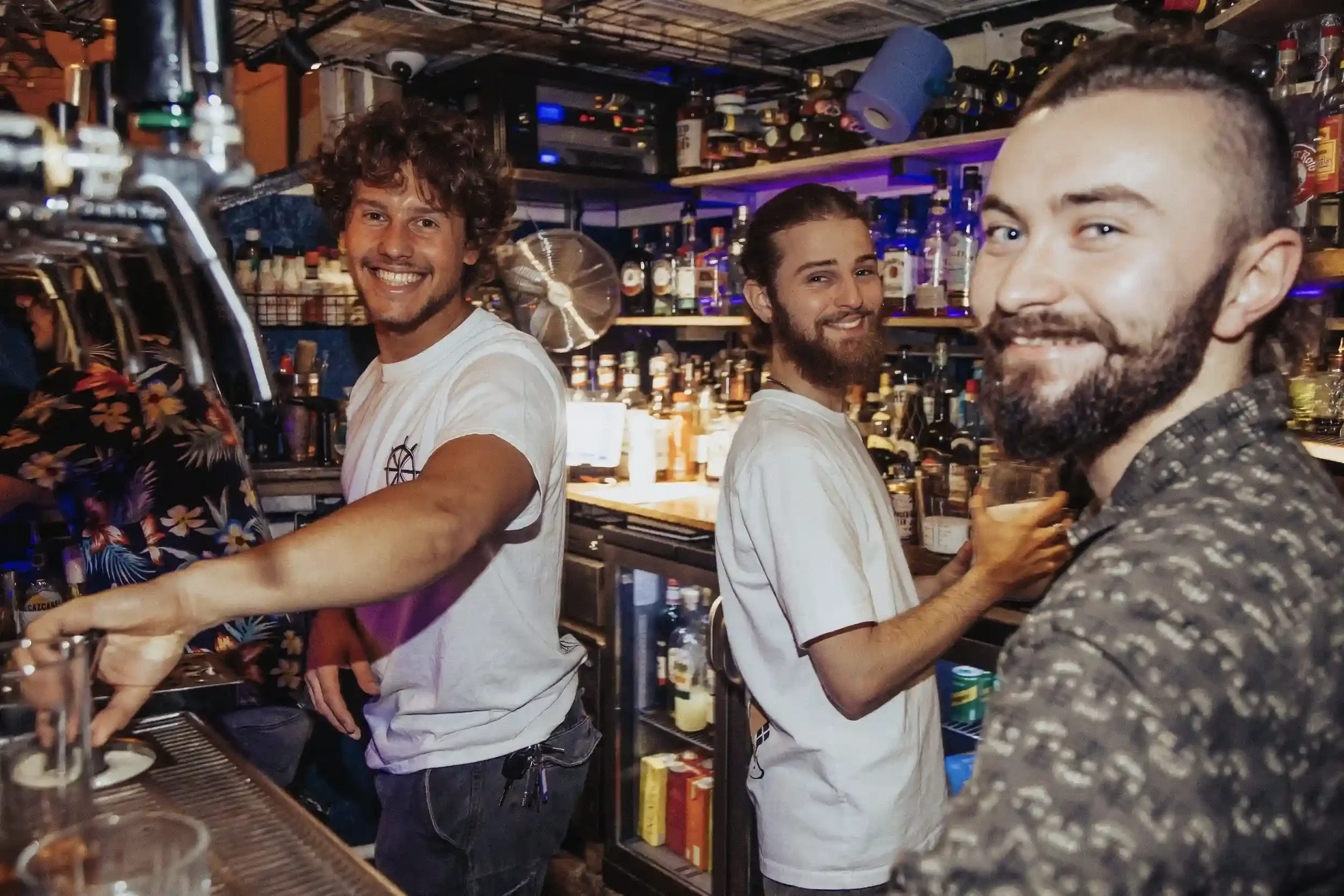 A high-contrast, candid photograph of three smiling bar staff members behind the bar at Small Ships. The staff are wearing white t-shirts, and the background is filled with a variety of spirits on shelves and beer taps. The direct flash lighting crea
