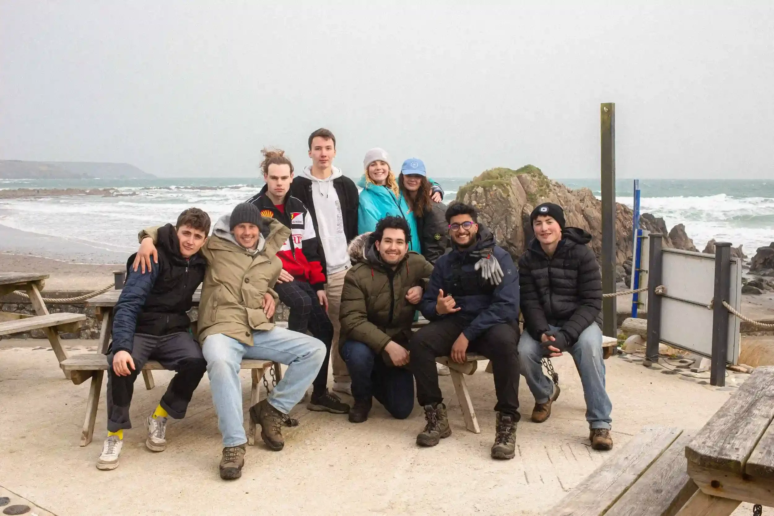 A behind-the-scenes colour group portrait of the 'Lighter' film cast and crew sitting and standing around a wooden picnic table on a concrete patio. The team is dressed in warm outdoor gear, with a rugged Cornish beach and white-capped sea in the bac
