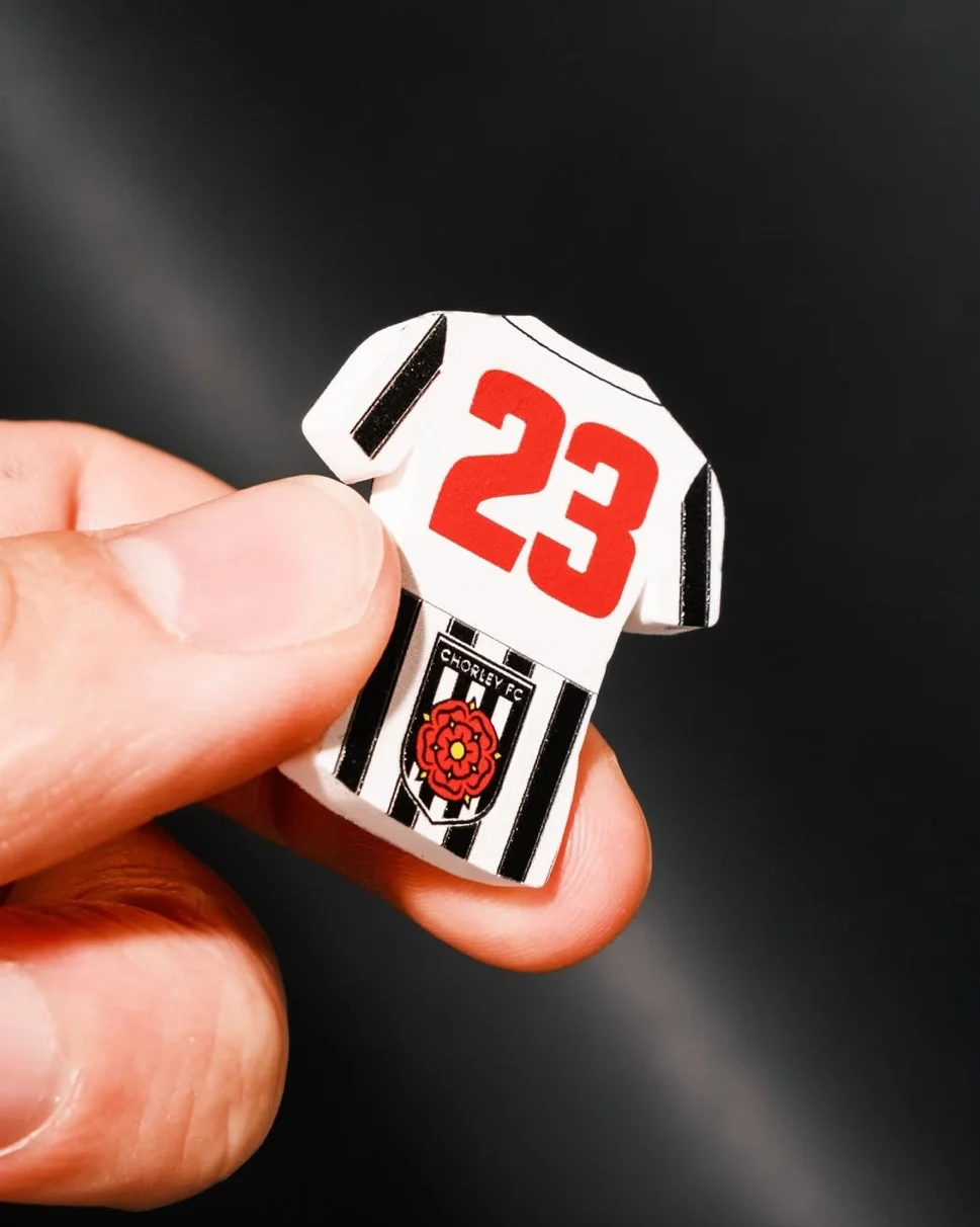 A macro close-up of a small, jersey-shaped magnet held in a person's hand. The magnet represents a Chorley FC kit with vertical black and white stripes on the lower half and a white upper half featuring a large red number '23'. The official Chorley F