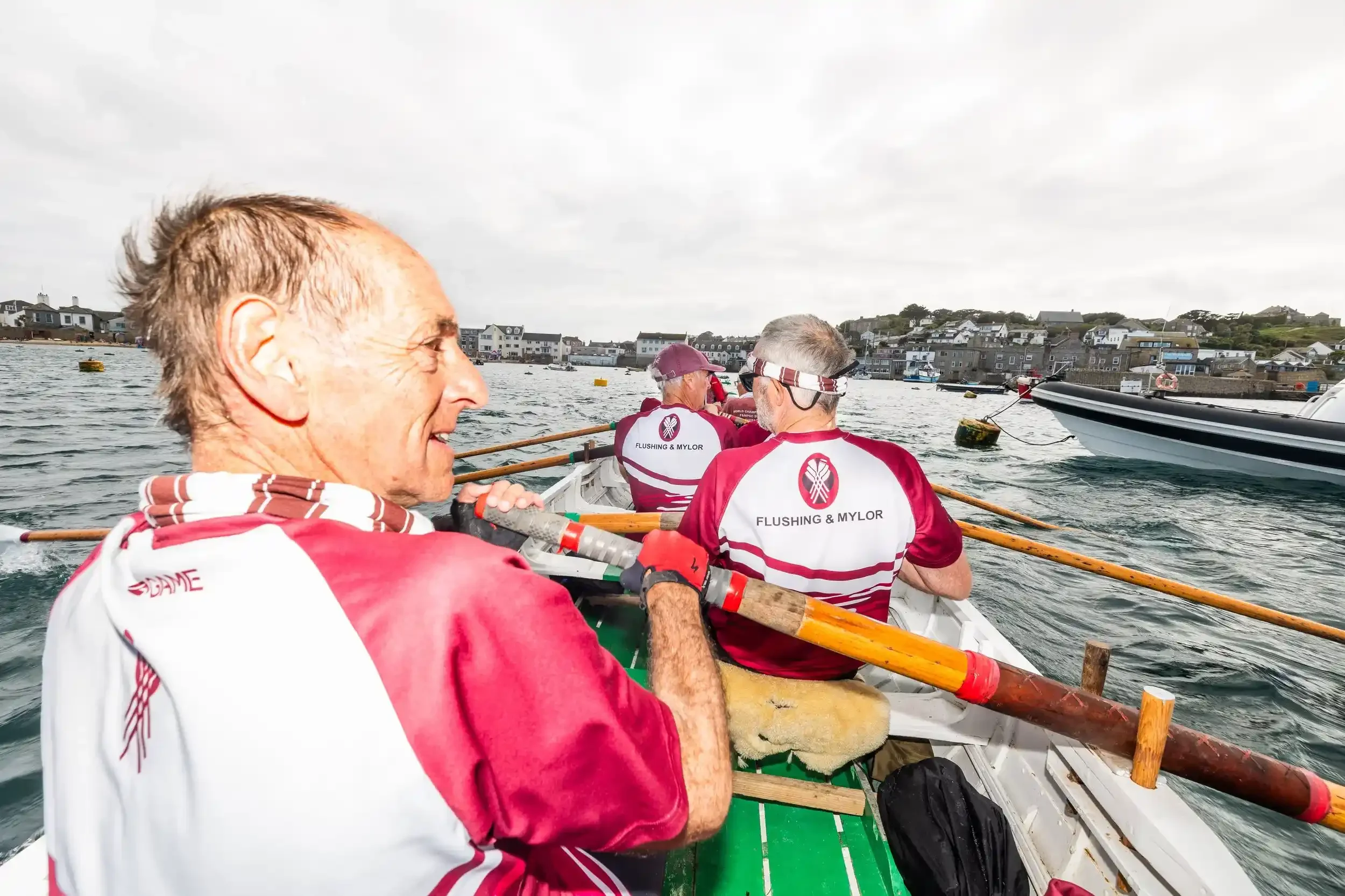 A first-person point-of-view shot from inside a wooden pilot gig during a race. In the foreground, the side profile of a man rowing is visible, his hair windswept. Looking down the length of the boat, two other rowers are seen from behind, wearing ma