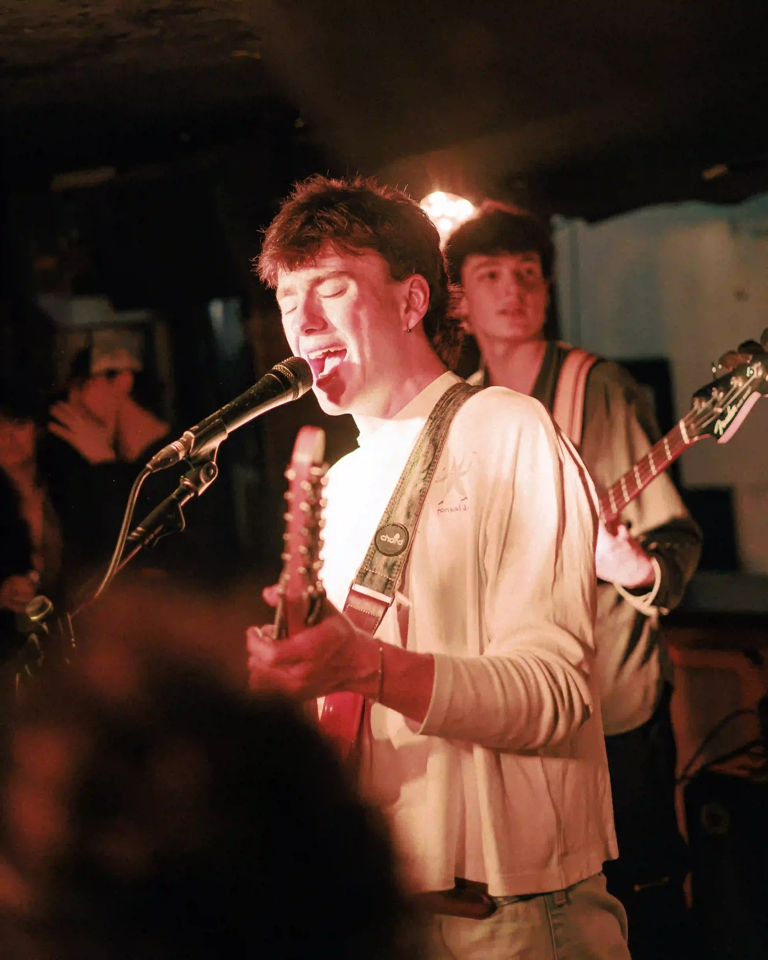 A vertical live music photograph of Tom Scamell singing with deep emotion at The Troubadour in London. He is captured from the chest up, wearing a white long-sleeve shirt with his eyes closed as he leans into a microphone. In the background, bassist 