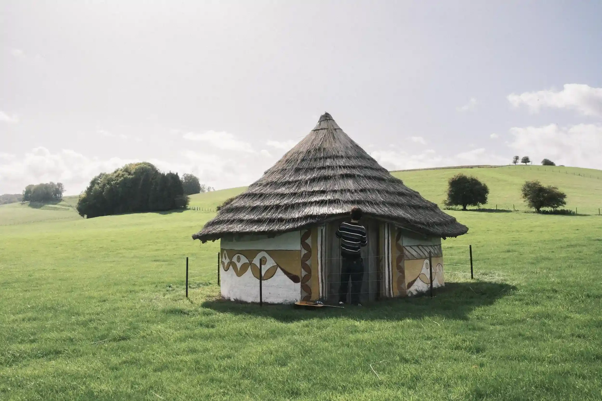 A wide-angle landscape photograph of musician Ned Holland standing in front of a small, circular hut with a high, pointed thatched roof. The hut's white walls are decorated with yellow and brown floral patterns and geometric borders. Holland stands w