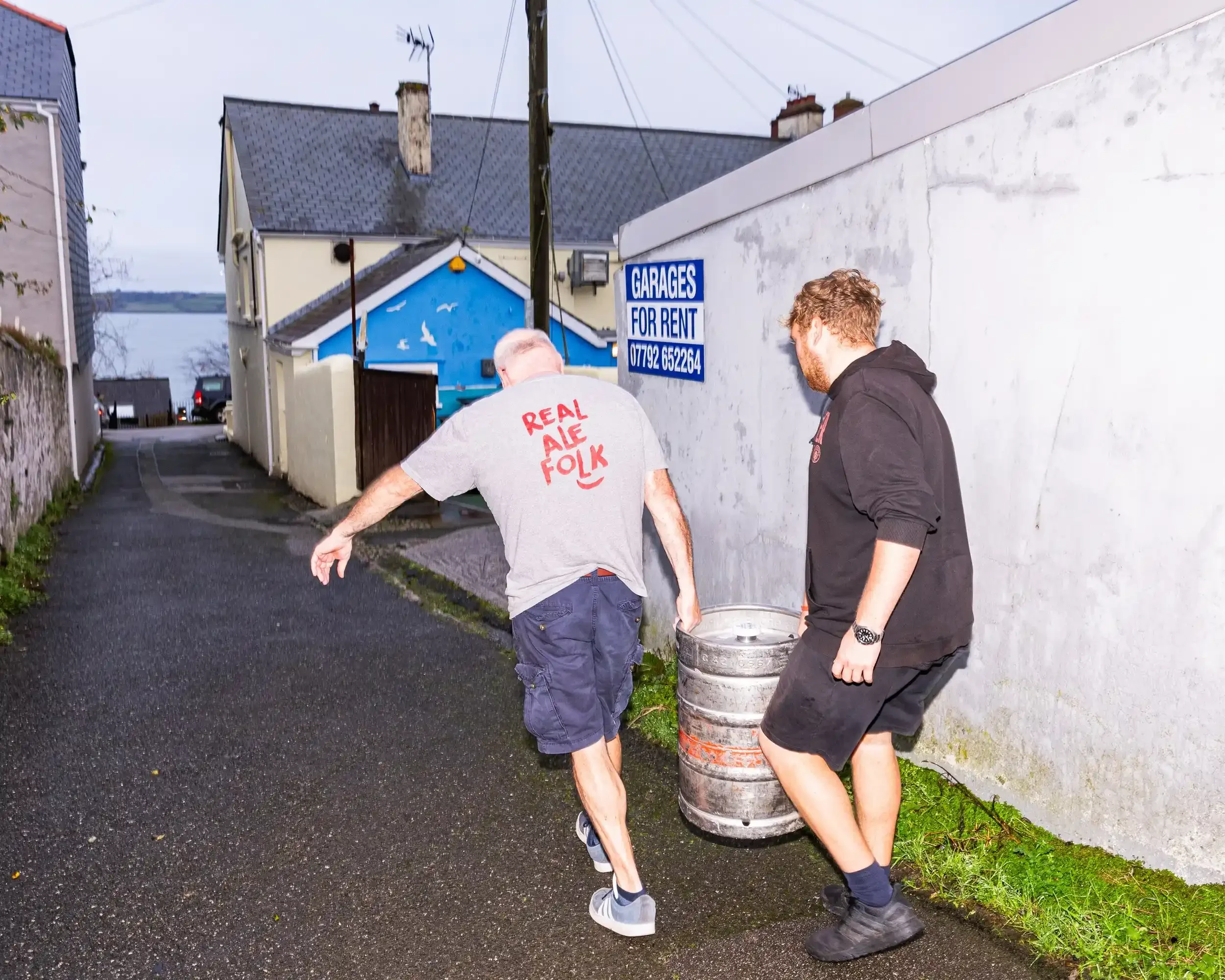 Two men carrying a heavy metal beer barrel up a narrow Falmouth street towards the Seaview Inn, by Matthew Morgan.