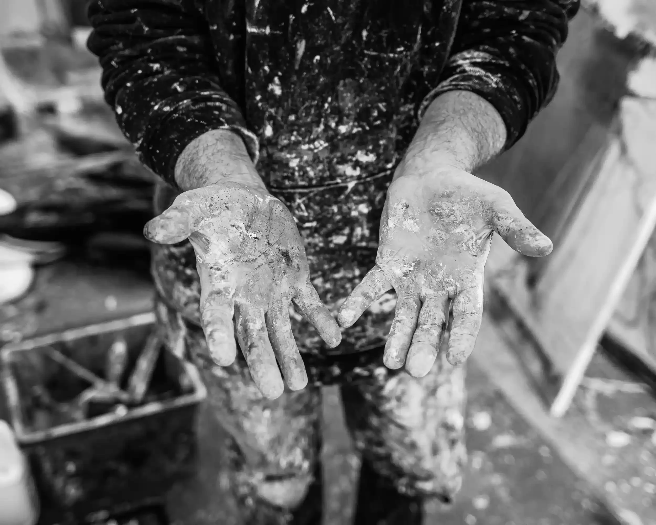 A stark black-and-white close-up of an artist's open palms, heavily coated in dried, cracked paint. The hands are held out toward the camera, emphasizing the thick layers of pigment on the skin. The artist, David Mankin, wears a dark, paint-splattere