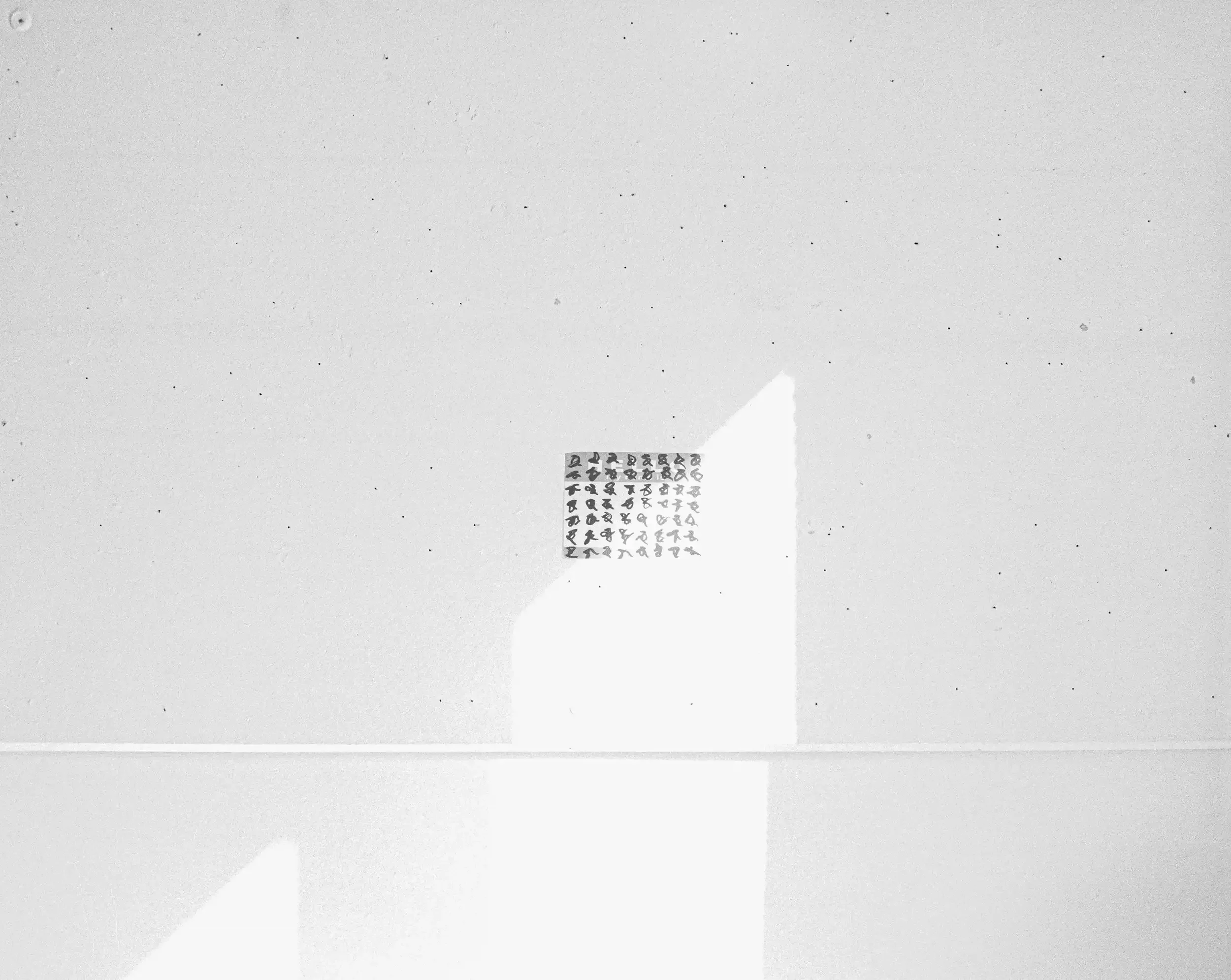 A minimalist black-and-white photograph of a small, square sheet of stickers featuring a grid of hand-drawn cryptic symbols. The sheet is stuck to a plain, textured white wall. A sharp, geometric shadow and a bright patch of sunlight cut across the w