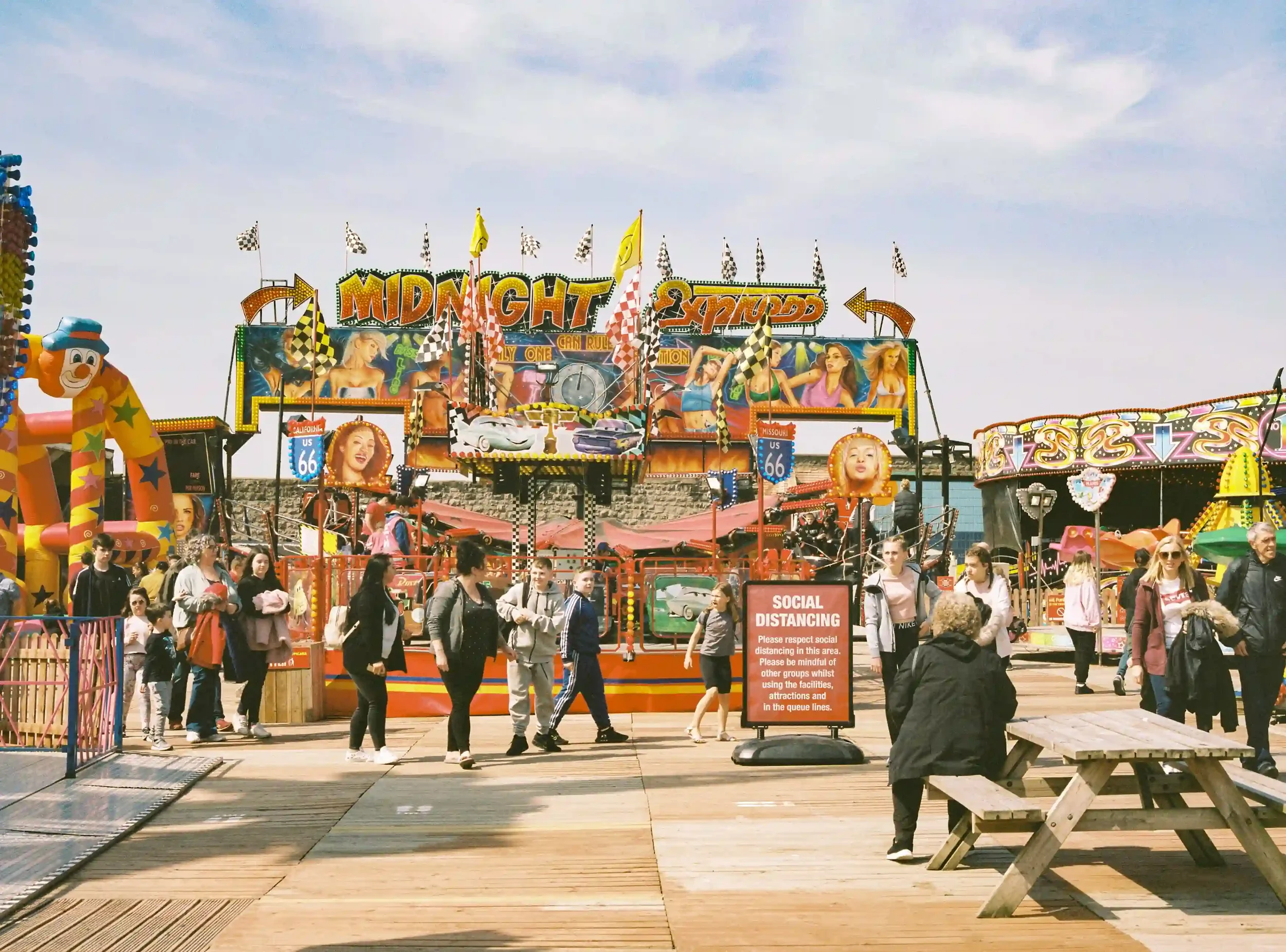 A busy funfair at Weston-super-Mare with colorful rides like the 'Midnight Express' in the background and a large red 'SOCIAL DISTANCING' sign in the foreground as people walk past on a wooden pier, by Matthew Morgan.