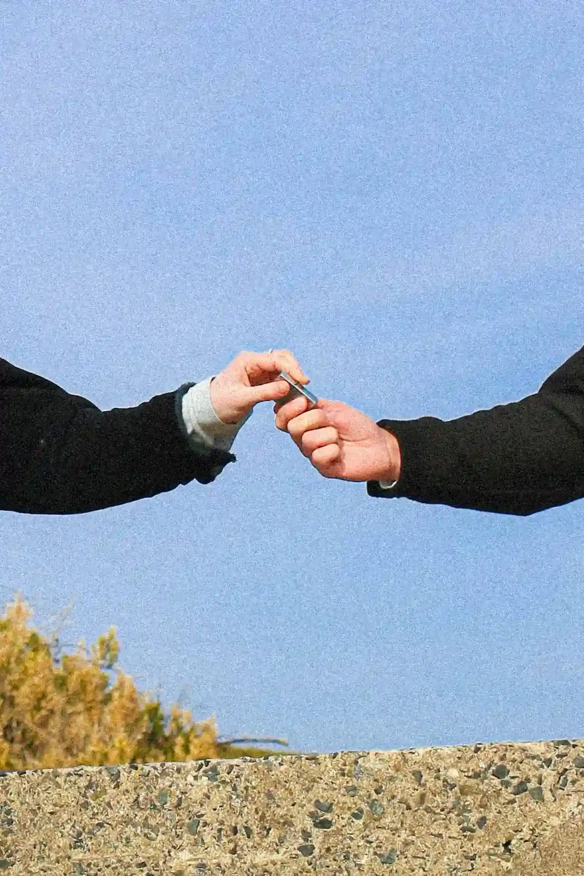 A cinematic colour unit still of two people's hands meeting in the center of the frame to exchange a small silver lighter. The arms are clad in dark sleeves and are set against a bright, grainy blue sky. Below the hands, a textured concrete wall and 