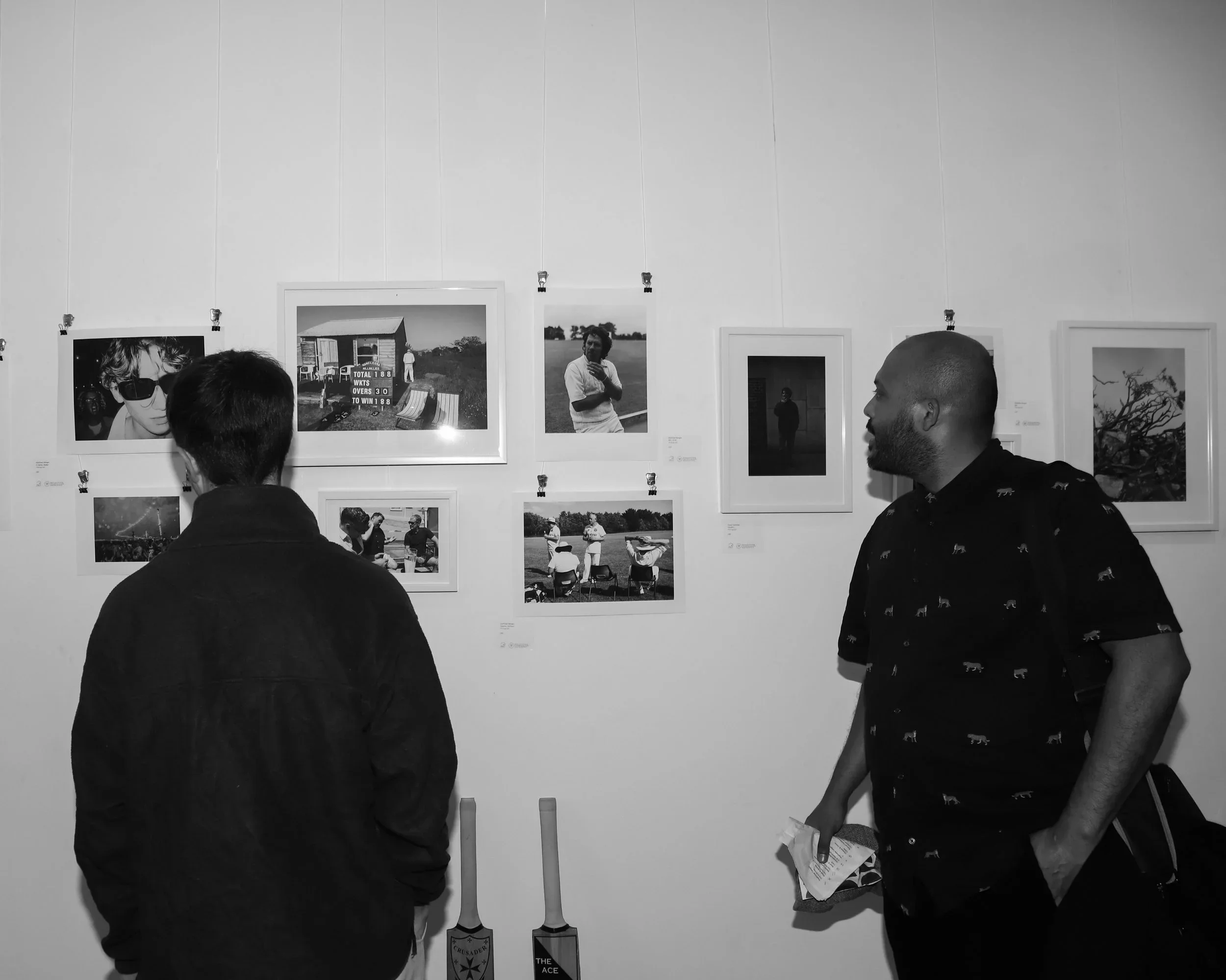 A black-and-white photograph of two men looking at a wall of art during an exhibition at The Poly in Falmouth. One man, seen from the back, is closely examining a series of photographs on the left. The other man, Wrihbu, is seen from the side, lookin