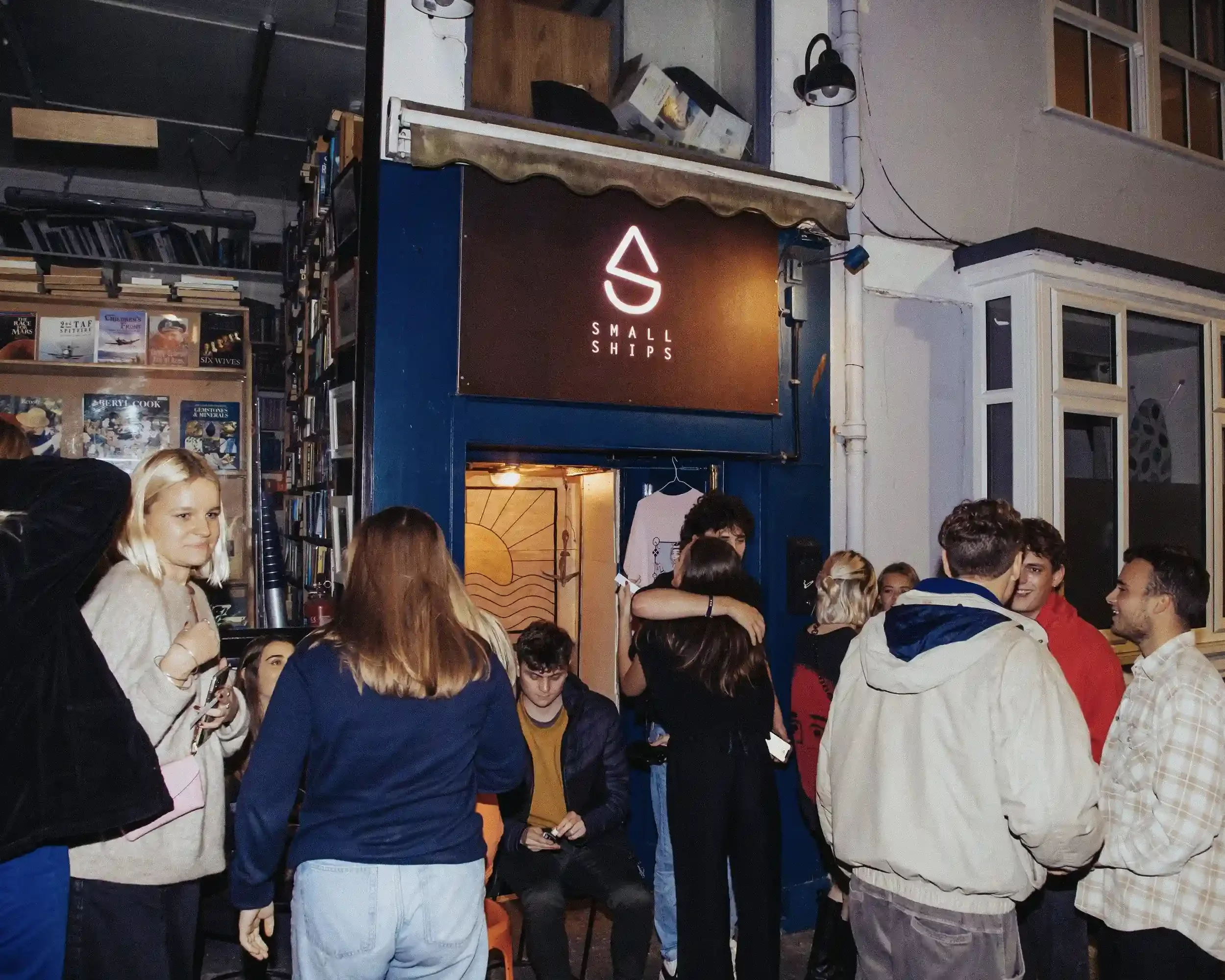 A night-time exterior shot of the Small Ships bar entrance. A dark brown sign with a glowing white neon logo and 'SMALL SHIPS' text sits above a doorway where people are hugging and socializing. To the left, a window reveals a room packed with books 