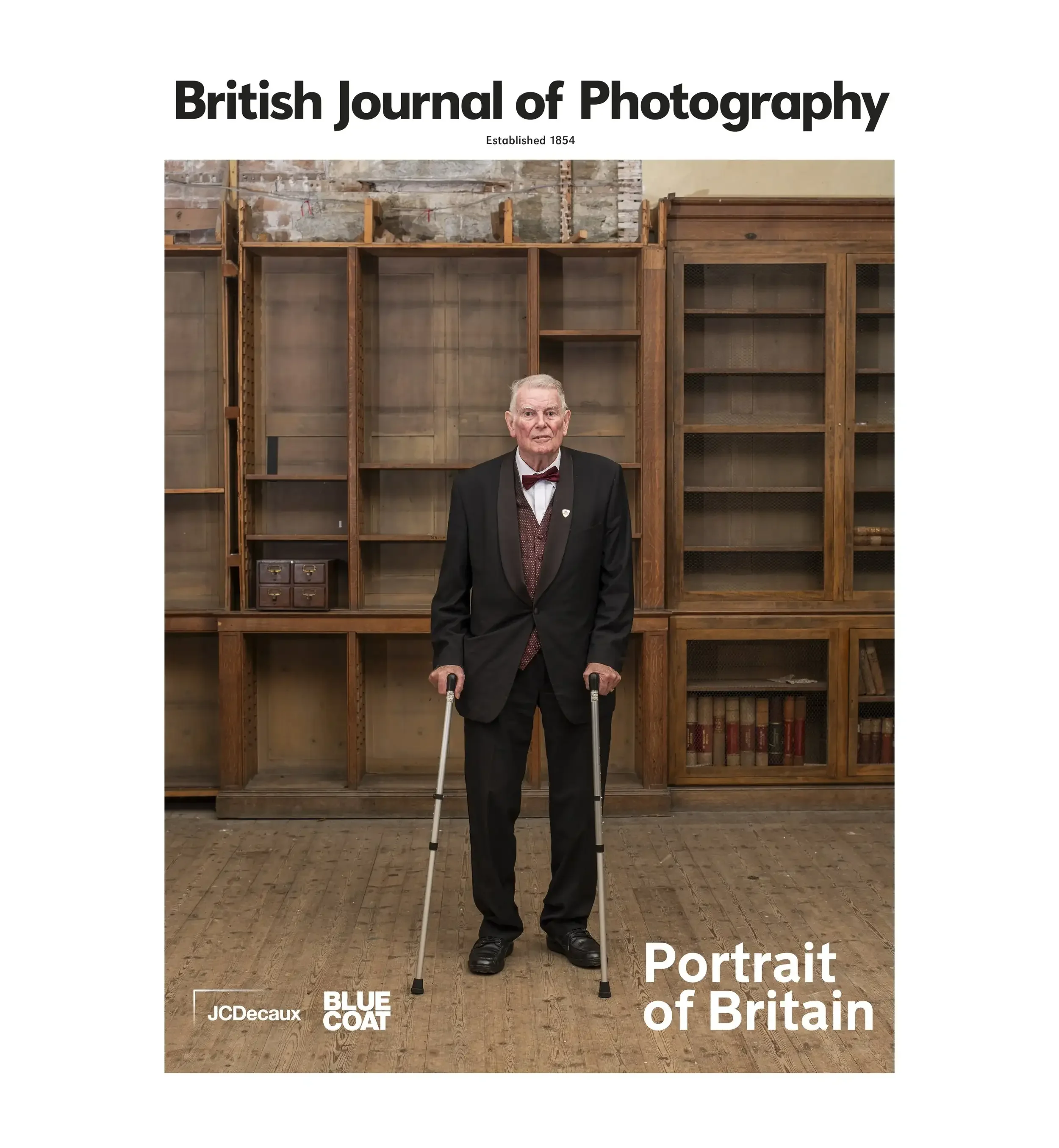 Official shortlisted portrait for the British Journal of Photography's Portrait of Britain. The image shows Peter Bray, an elderly man in a black tuxedo and red bow tie, standing with crutches in a room with vintage wooden library shelves. The 'Briti