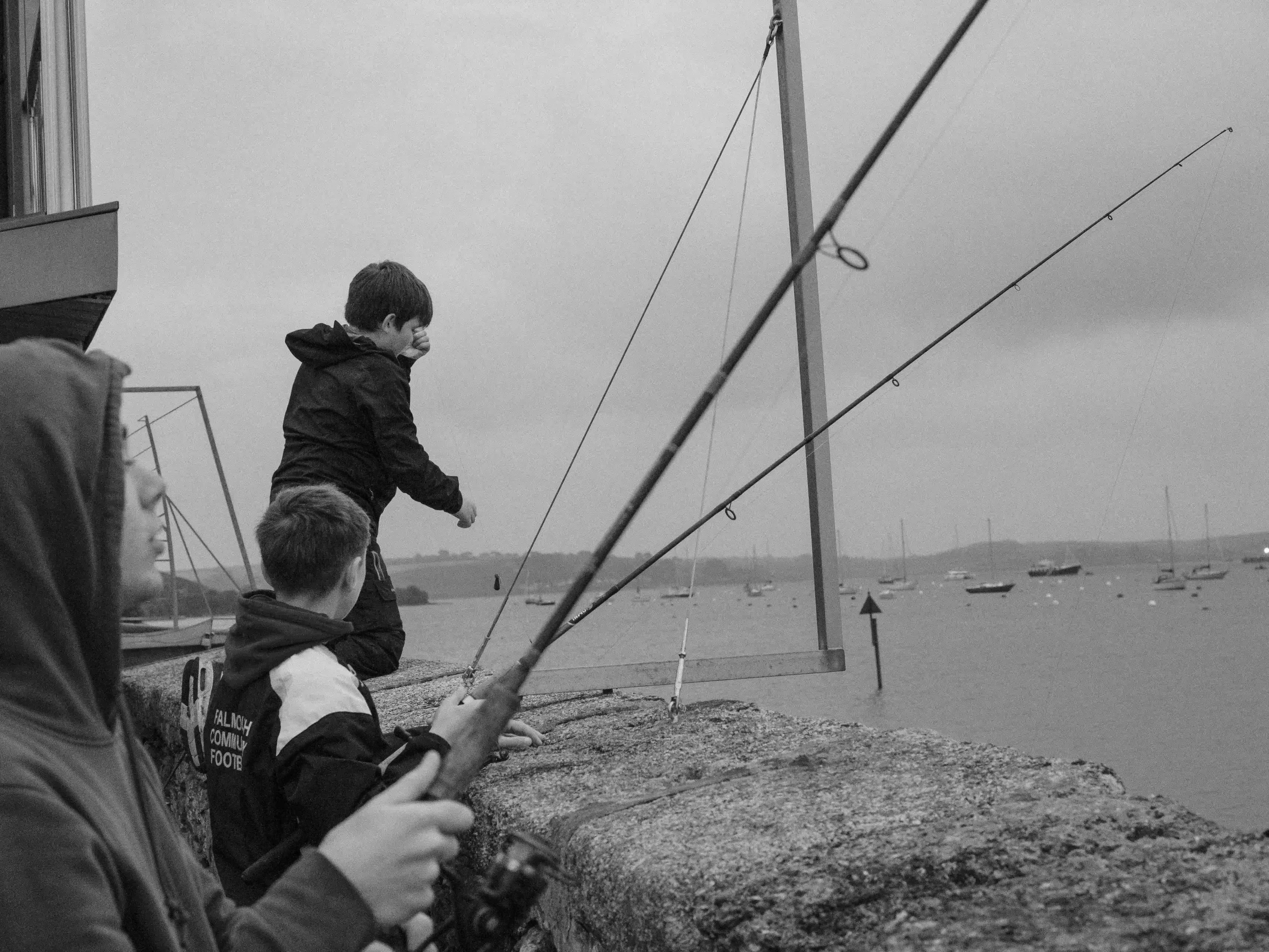 A black-and-white photograph of three young people fishing from a stone harbor wall in Flushing. Two long fishing rods are silhouetted against a cloudy sky, pointing toward a harbor filled with sailing boats, while one boy in a dark hoodie wipes his 