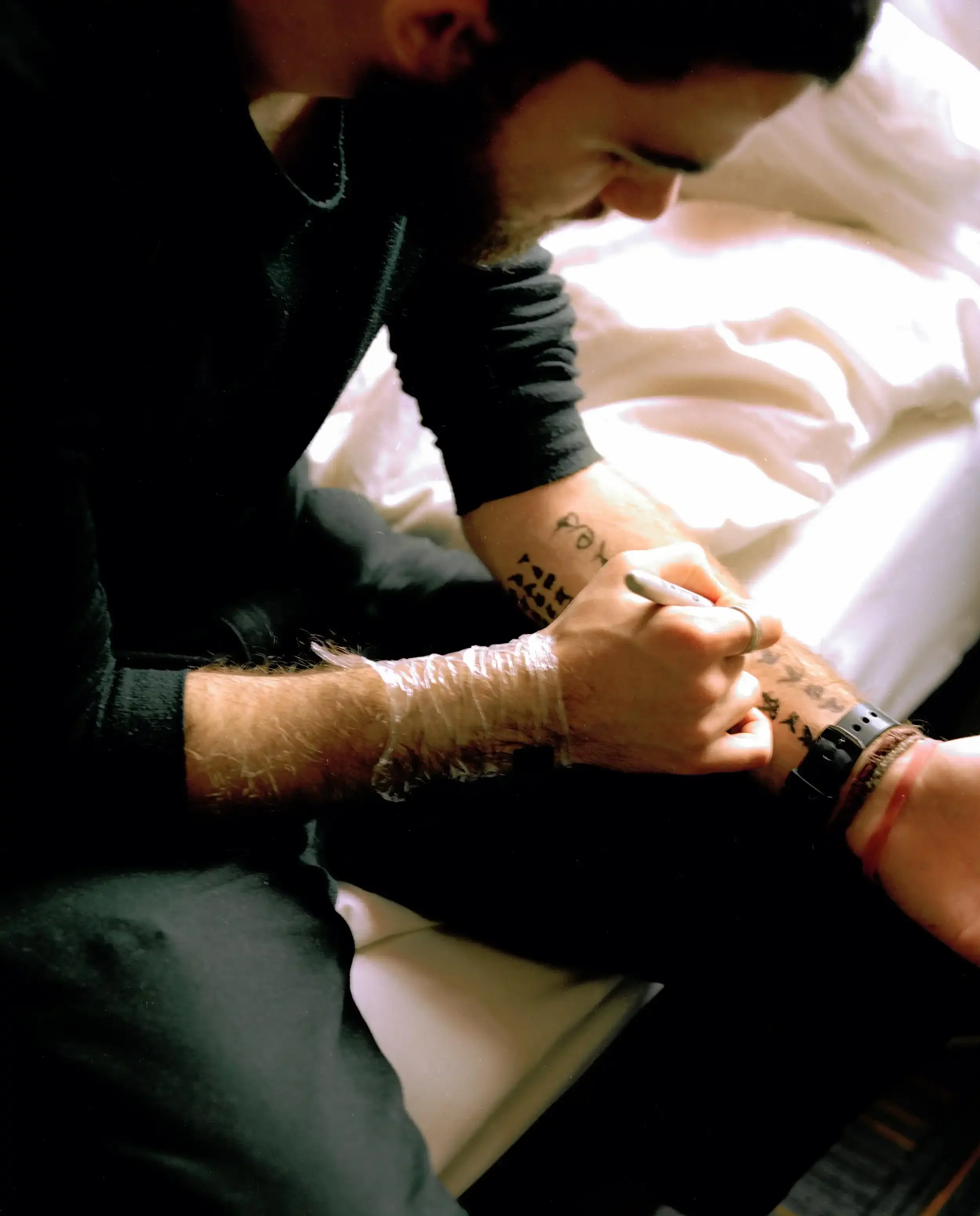 A close-up, high-contrast photograph of artist Finlay Cummins sitting on a white bed, leaning over to draw intricate black symbols on his own forearm with a pen. His other wrist is wrapped in clear plastic, and he wears a dark long-sleeved shirt. The