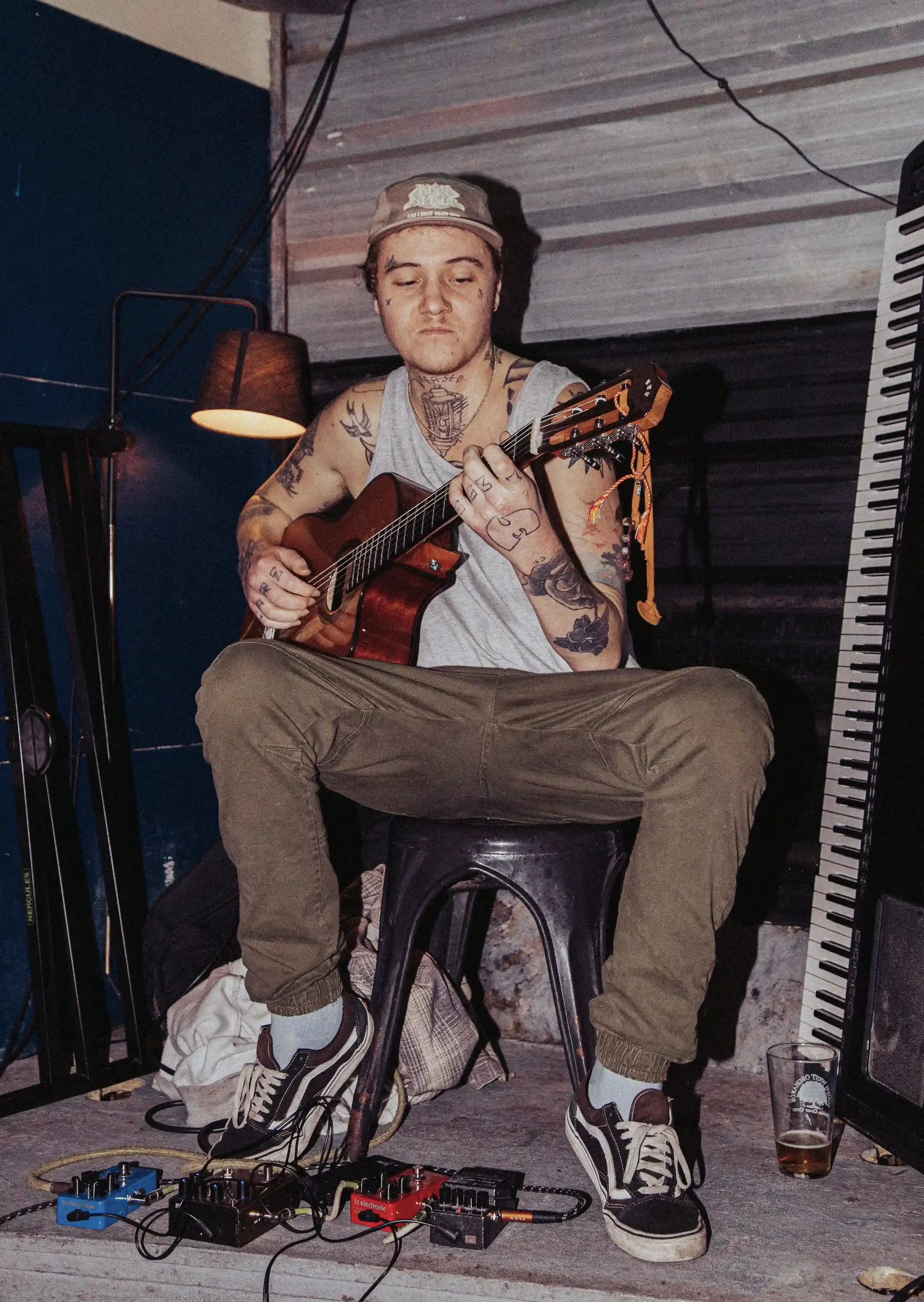 A gritty, high-contrast portrait of a musician sitting on a stool and playing an acoustic guitar inside Small Ships bar. He is wearing a grey vest, khaki trousers, and a cap, with visible tattoos on his arms and hands. Several guitar pedals are on th