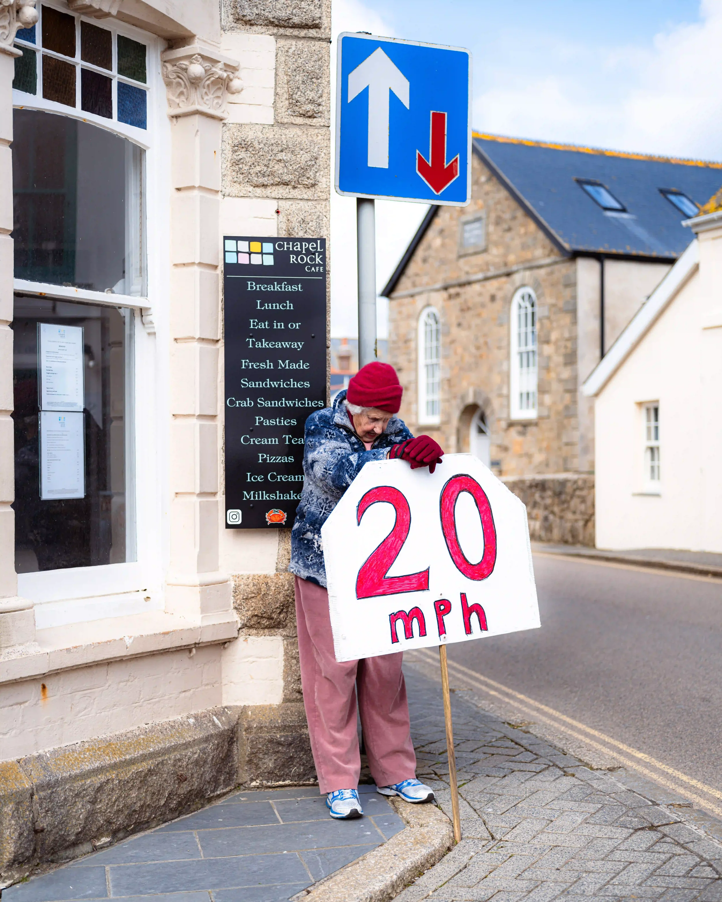 An elderly woman, Mrs. P, wearing a red beanie, blue patterned fleece, and pink trousers, leans against a large, hand-drawn '20 mph' sign on a wooden stake. She is standing on a cobbled corner next to the Chapel Rock Cafe in Marazion, Cornwall, under
