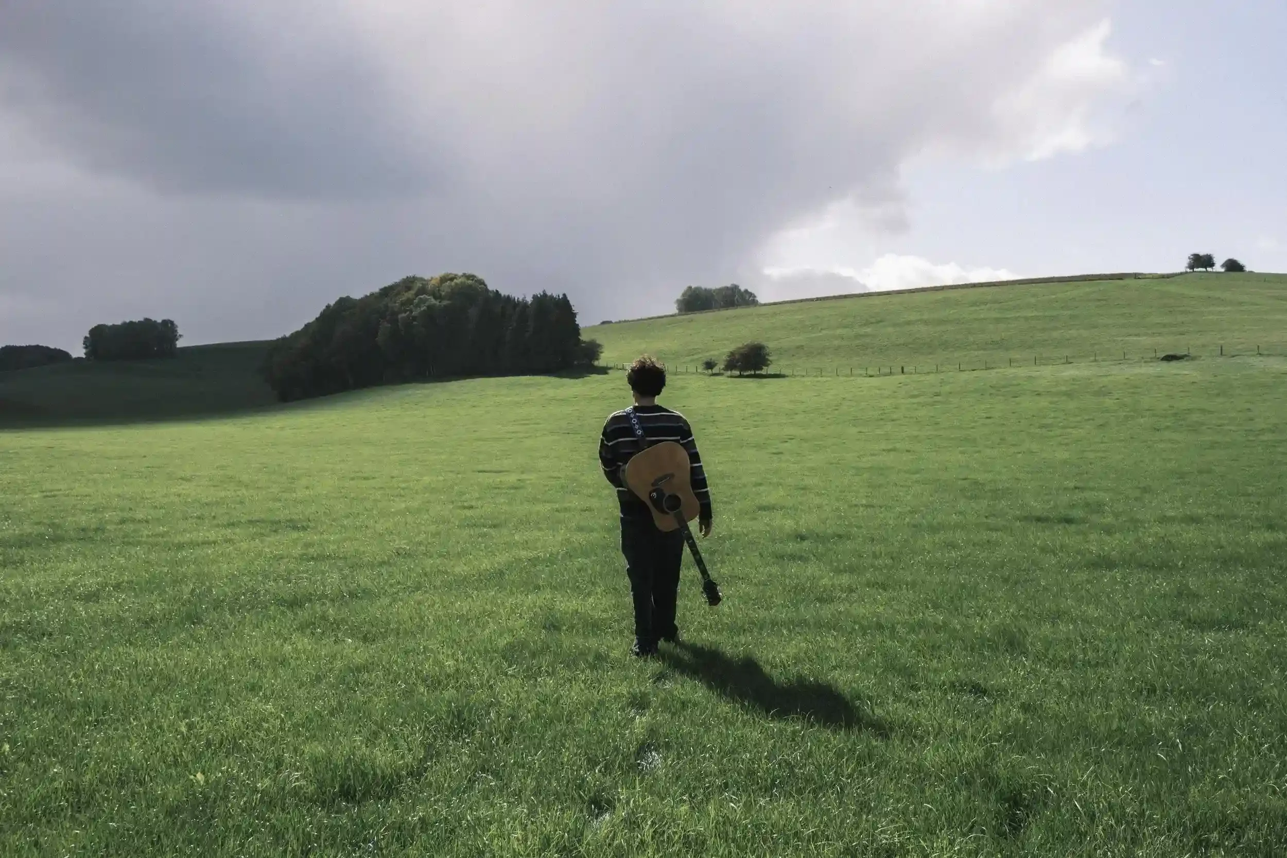 A wide-angle landscape photograph of musician Ned Holland walking away from the camera into a vast, sunlit green field in Salisbury. Holland wears a dark striped jumper and has an acoustic guitar strapped to his back. The field is bordered by thick g