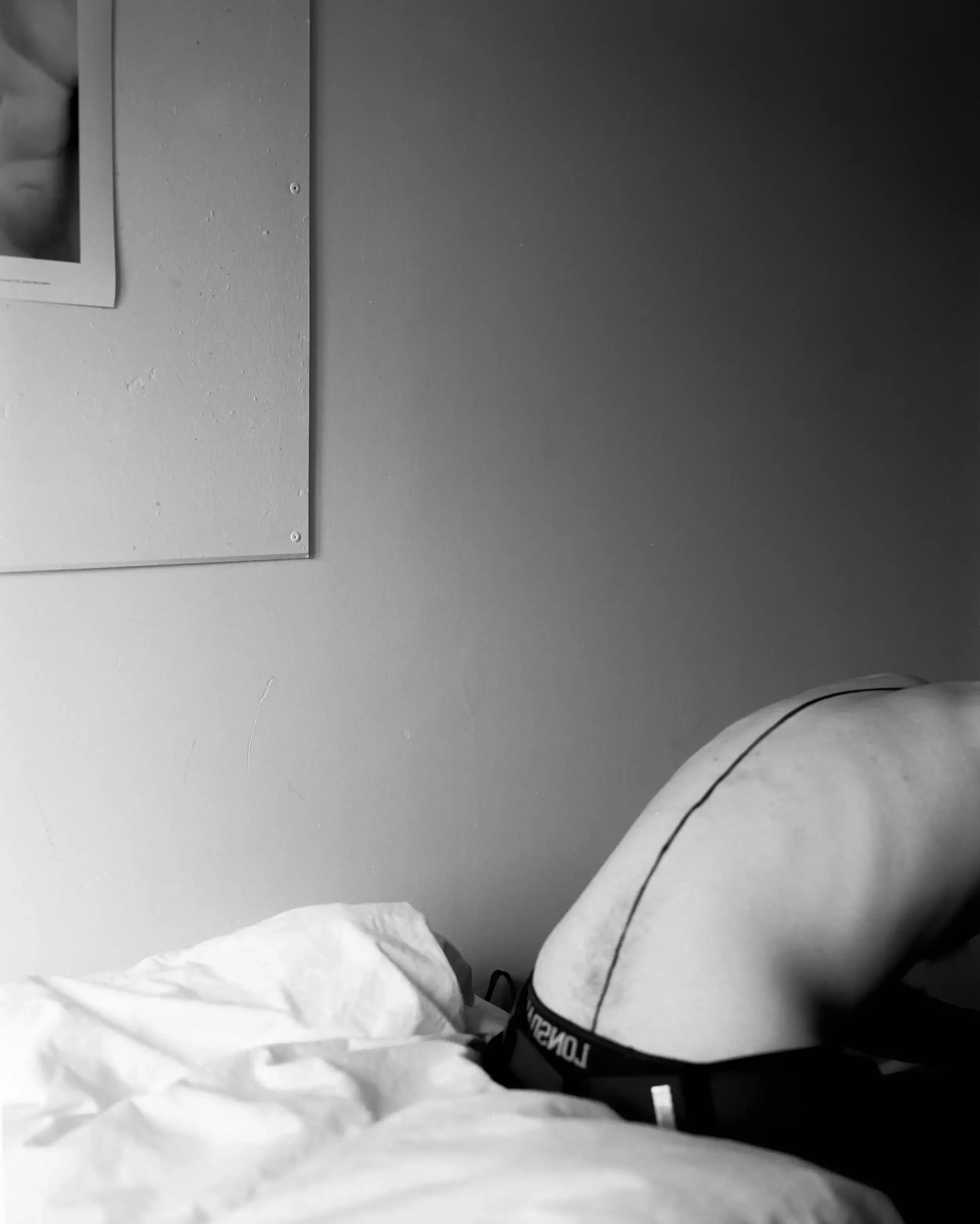 A minimalist black-and-white photograph of the bare back of artist Finlay Cummins, arched over a white bed. A single, dark, hand-drawn line runs vertically down the center of his spine, disappearing into the waistband of his dark trousers. A portion 