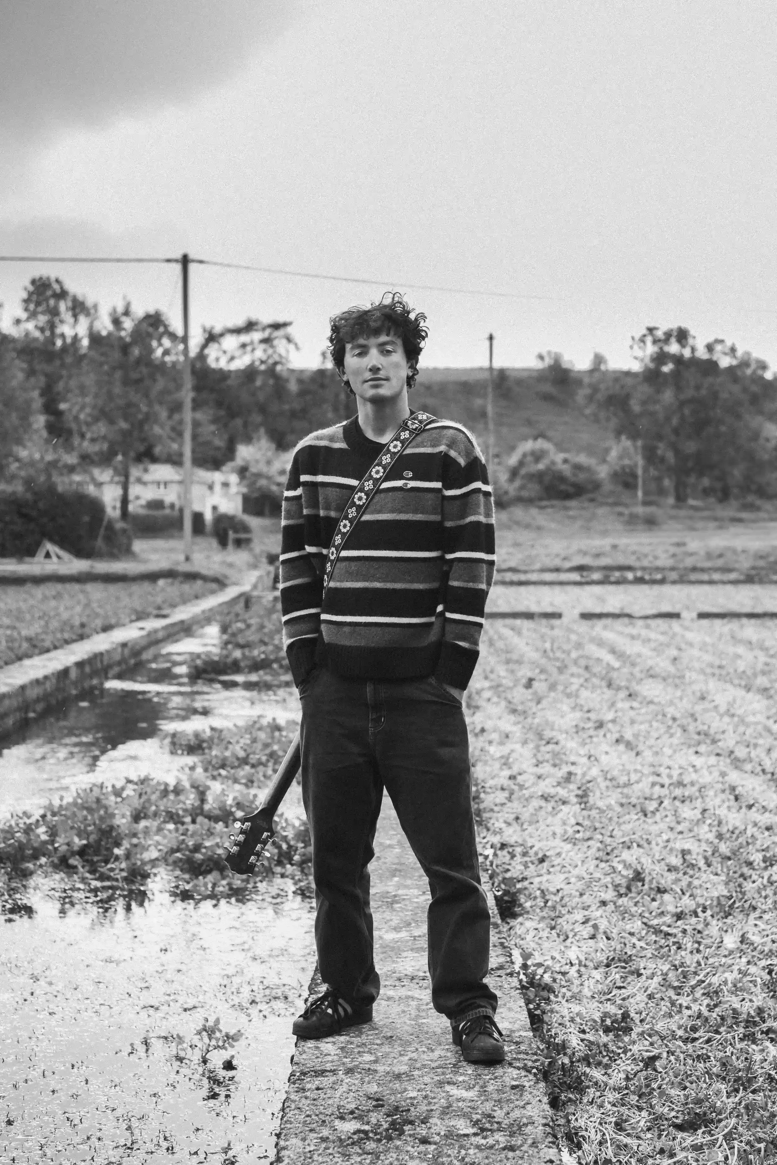 A black-and-white full-body portrait of musician Ned Holland standing on a low stone wall next to a flowing stream in a rural field. Holland is looking at the camera, wearing a dark striped jumper and jeans, with an acoustic guitar held by his side. 