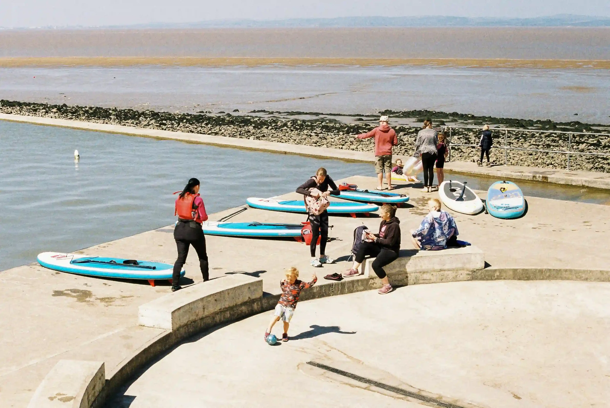A sunny day at Clevedon Marine Lake featuring people in wetsuits and casual clothes standing near several blue and white paddleboards on a concrete pier, with a child kicking a ball in the foreground and the muddy Severn Estuary in the background, by