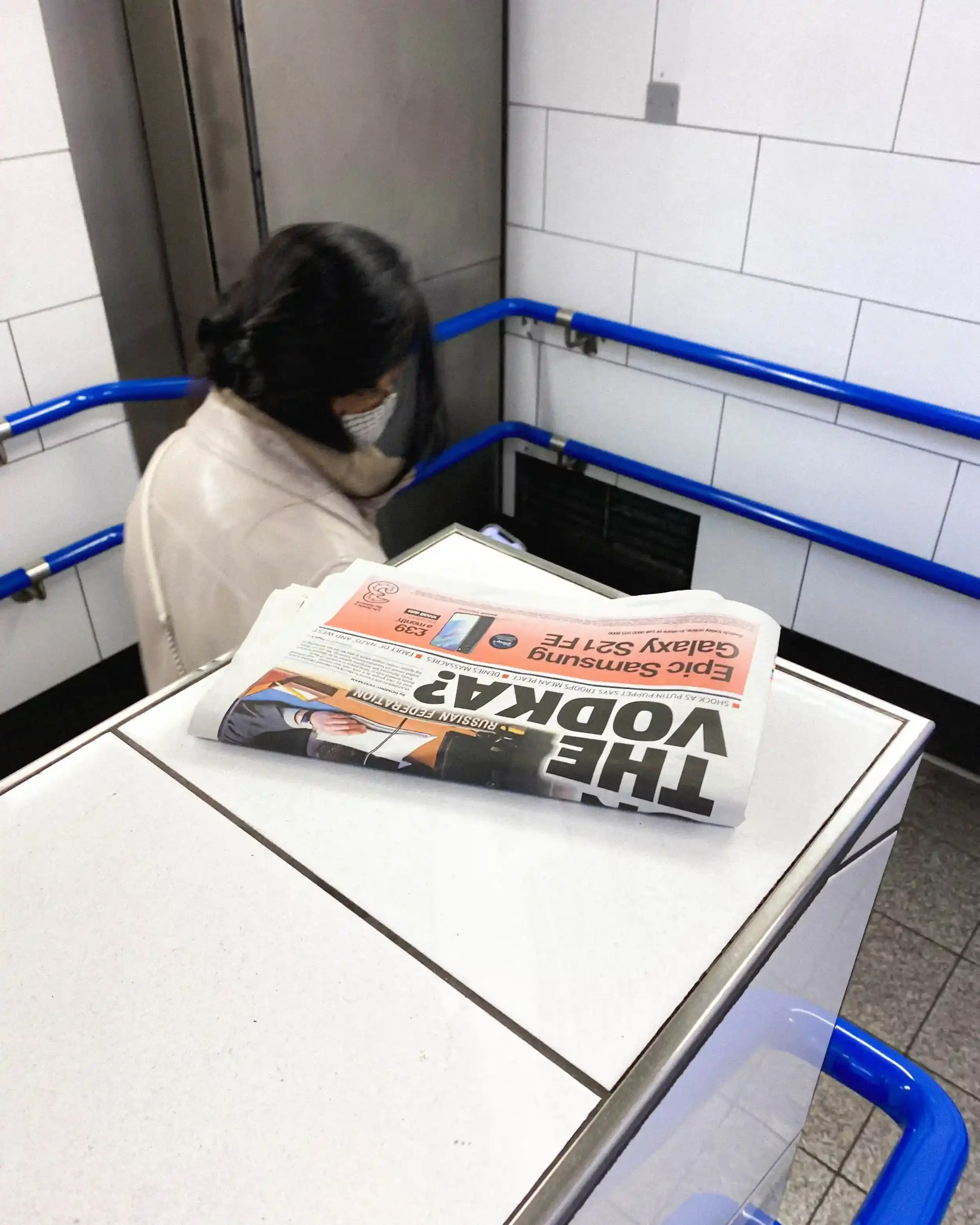 A discarded newspaper with a headline about 'The Vodka' resting on a ledge in a London Underground station as a commuter walks away, from the series 'London's Free Newspapers' by Matthew Morgan.