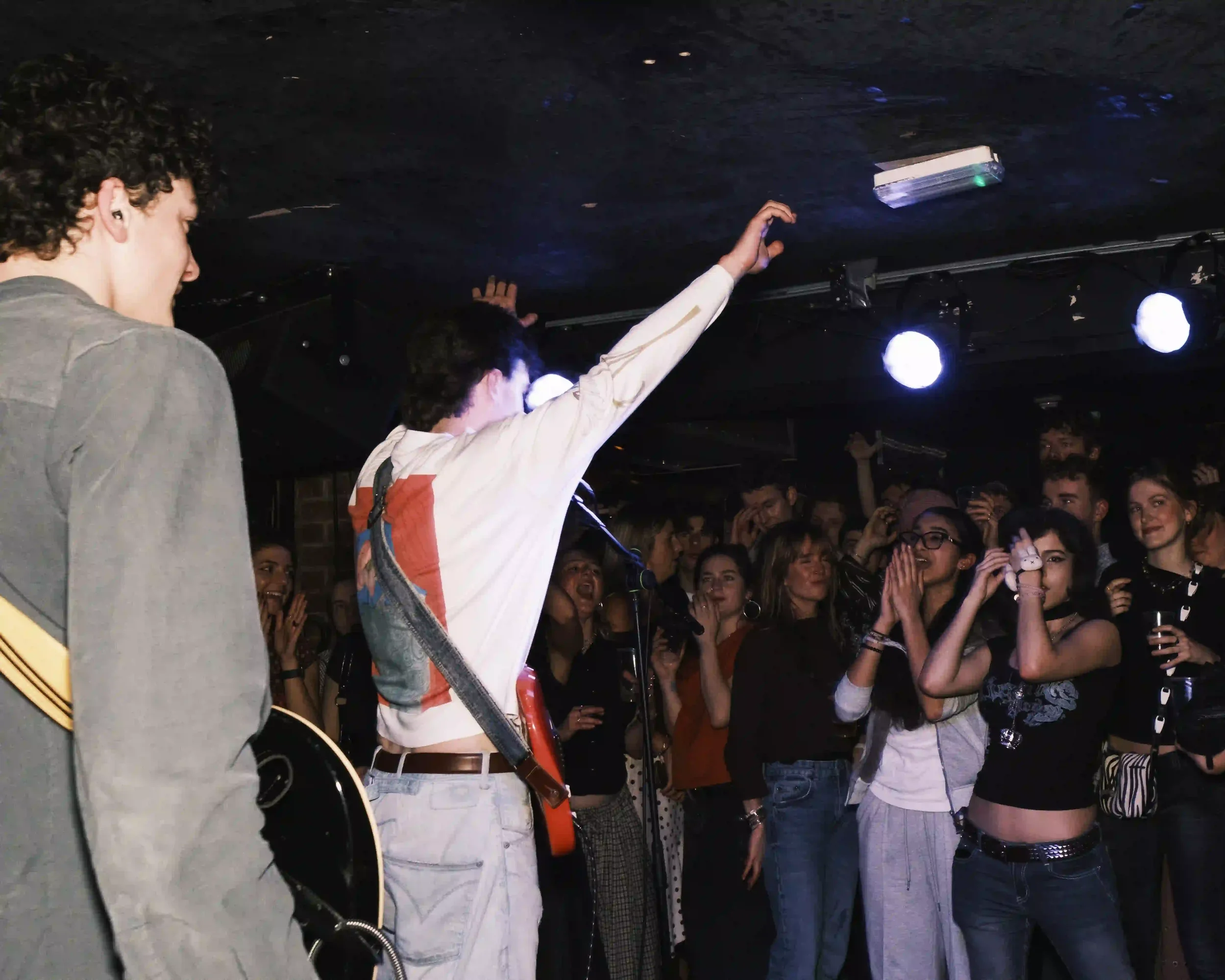 A live music photograph from behind the stage at The Troubadour, London. Tom Scamell, seen from the back in a white long-sleeve shirt and light jeans with a red guitar strapped on, raises his right arm to thank a cheering crowd. A sea of fans in the 