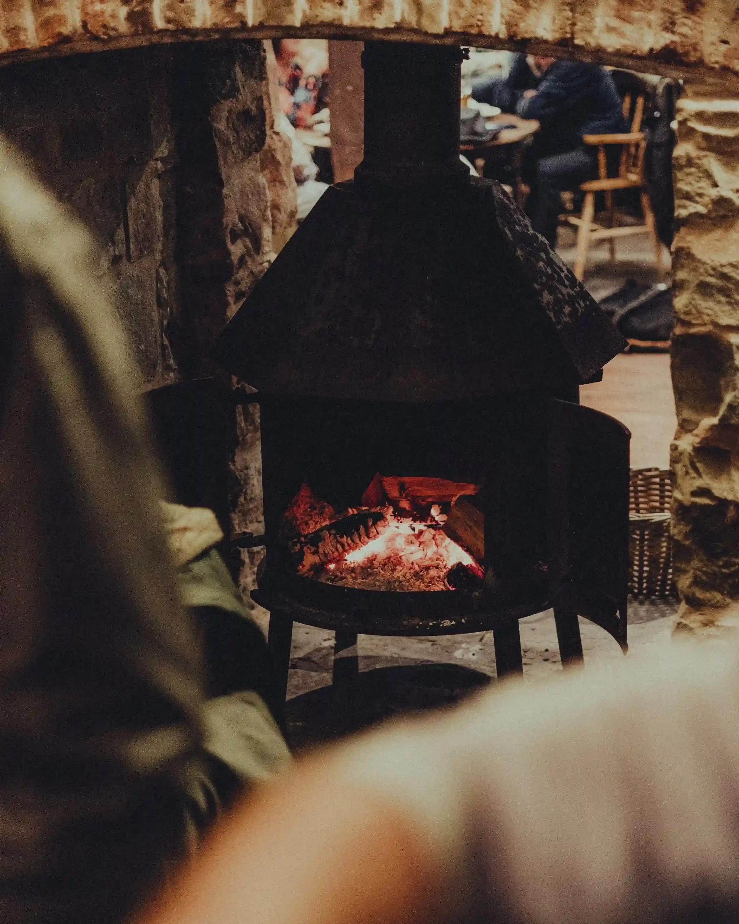 A warm, inviting interior shot of a log-burning stove inside a rustic brick fireplace at The Crown Inn, Lea. Bright orange flames are visible through the glass doors of the black metal stove. The foreground is slightly blurred, showing a wooden table