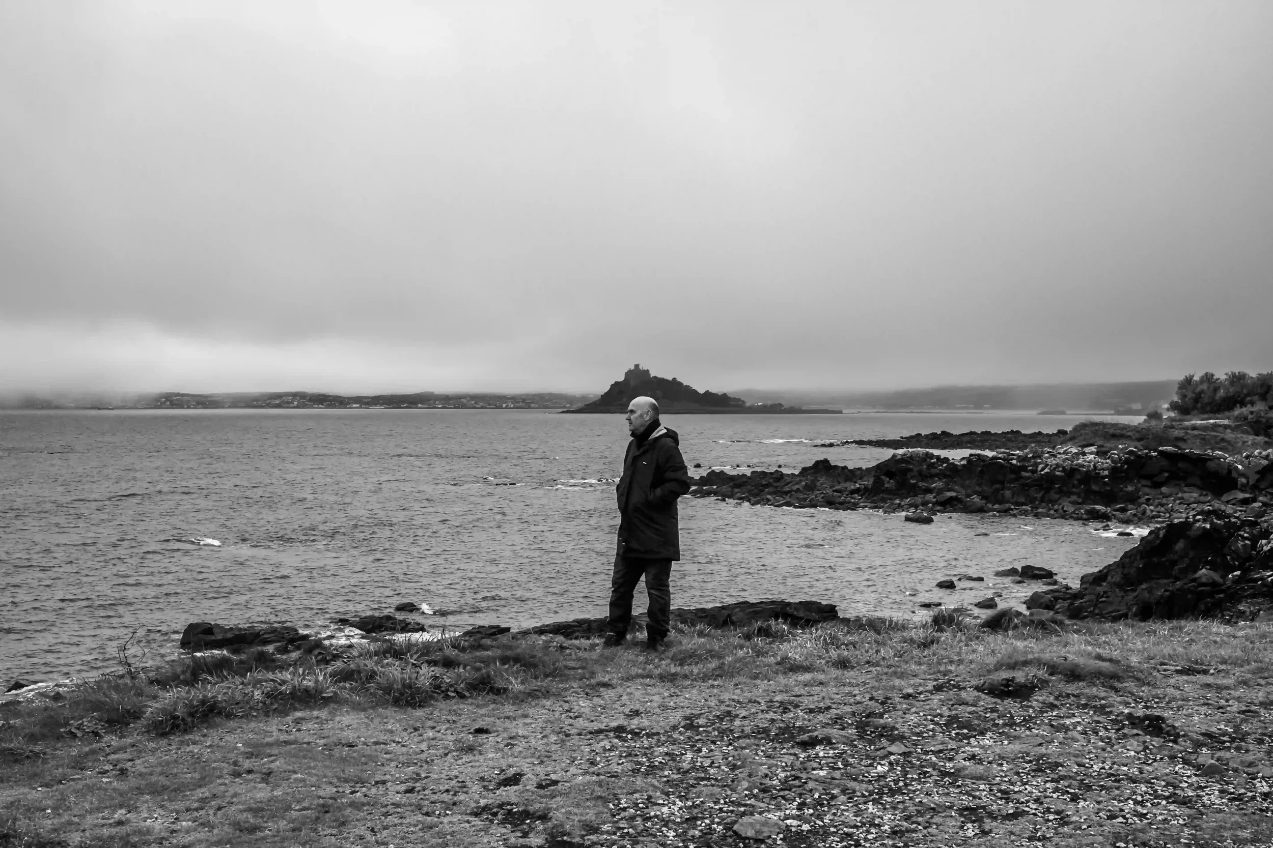 A wide-angle black-and-white environmental portrait of artist David Mankin standing on a grassy, rocky shoreline in Cornwall. He is wearing a dark winter coat with his hands in his pockets, looking out across the water. In the distance, the misty sil
