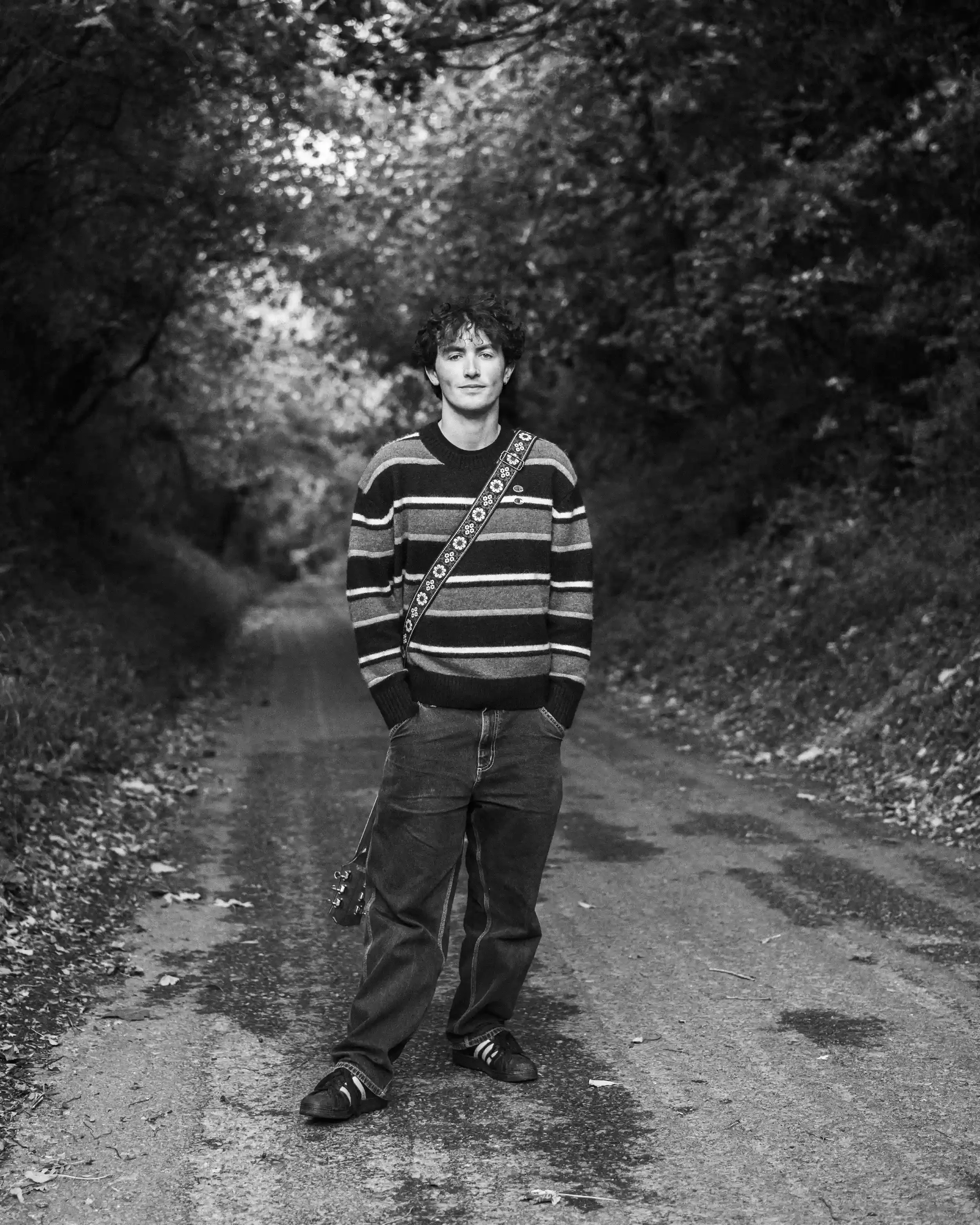 A black-and-white full-body portrait of musician Ned Holland for his single 'Bobbys.' Holland stands centrally on a narrow, tree-lined country lane, looking directly at the camera with a calm expression. He wears a dark, horizontally striped sweater 