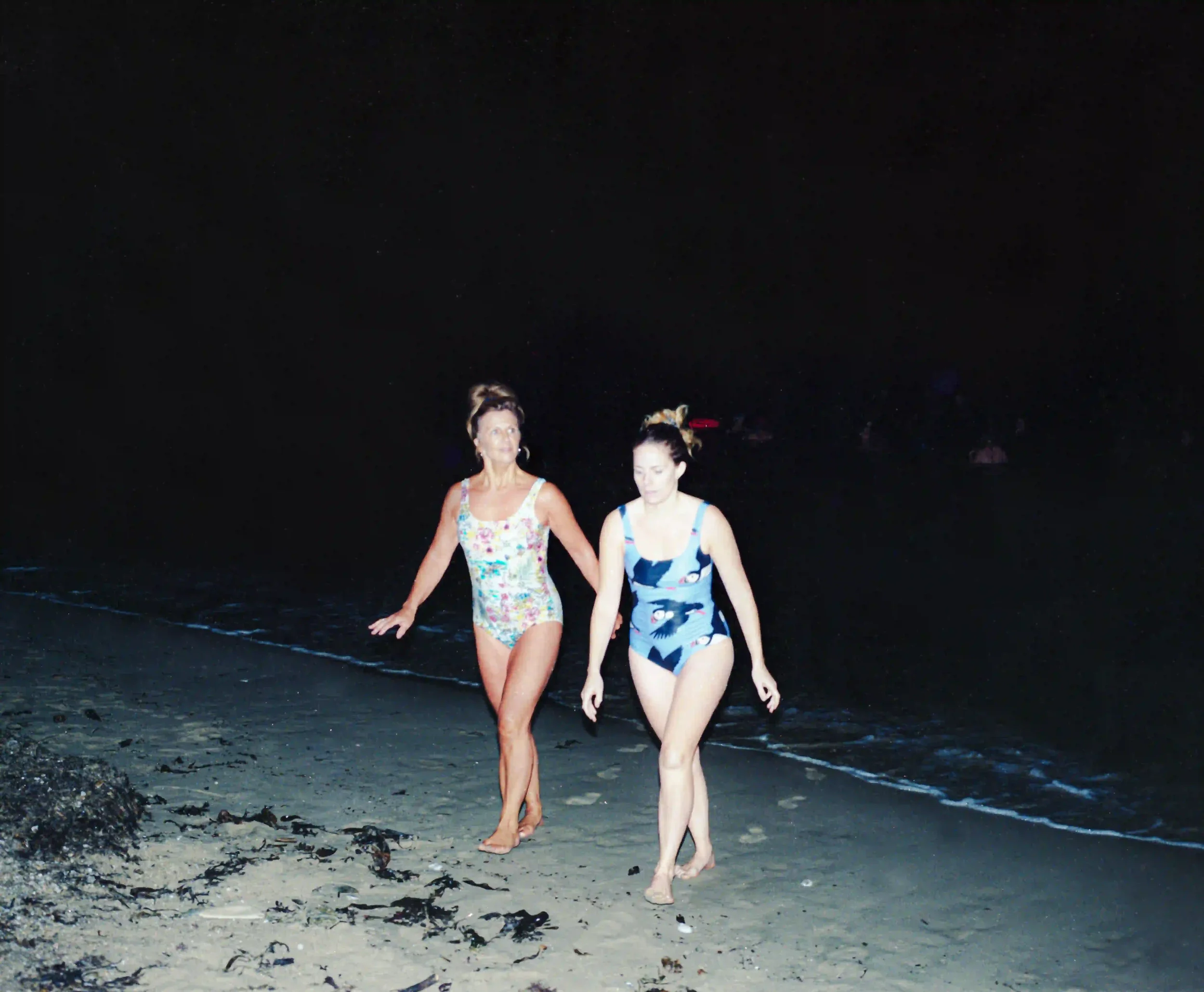 A night-time flash photograph of two women walking on the dark sands of Swanpool Beach, Falmouth, after a swim. One woman on the left wears a colorful floral swimsuit, while the woman on the right wears a blue swimsuit with a puffin pattern. The back