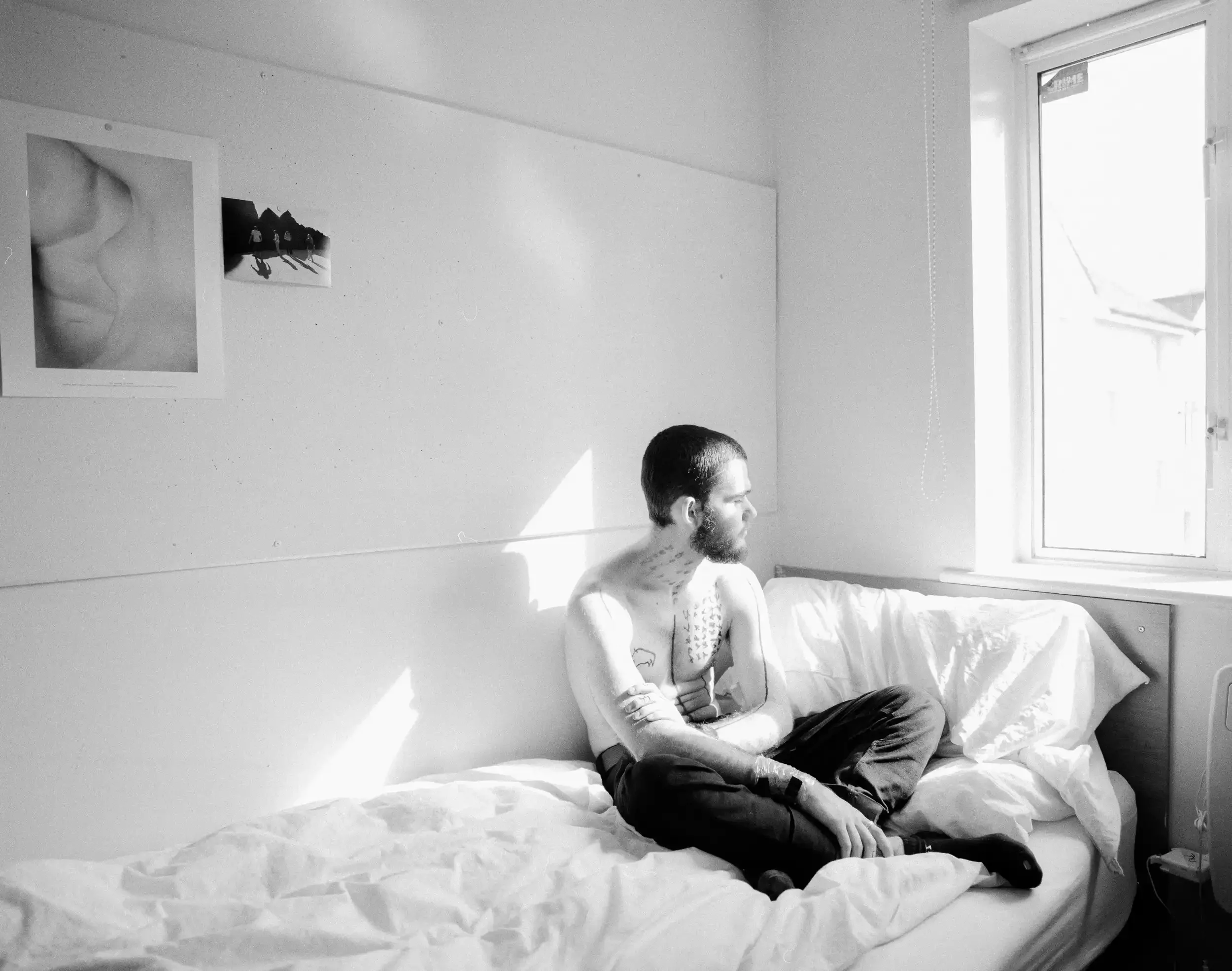 A wide-angle black-and-white photograph of a young man, artist Finlay Cummins, sitting cross-legged on a white unmade bed in a minimalist room. He is shirtless, showing extensive dark hand-drawn markings on his chest and arms, and is looking toward a