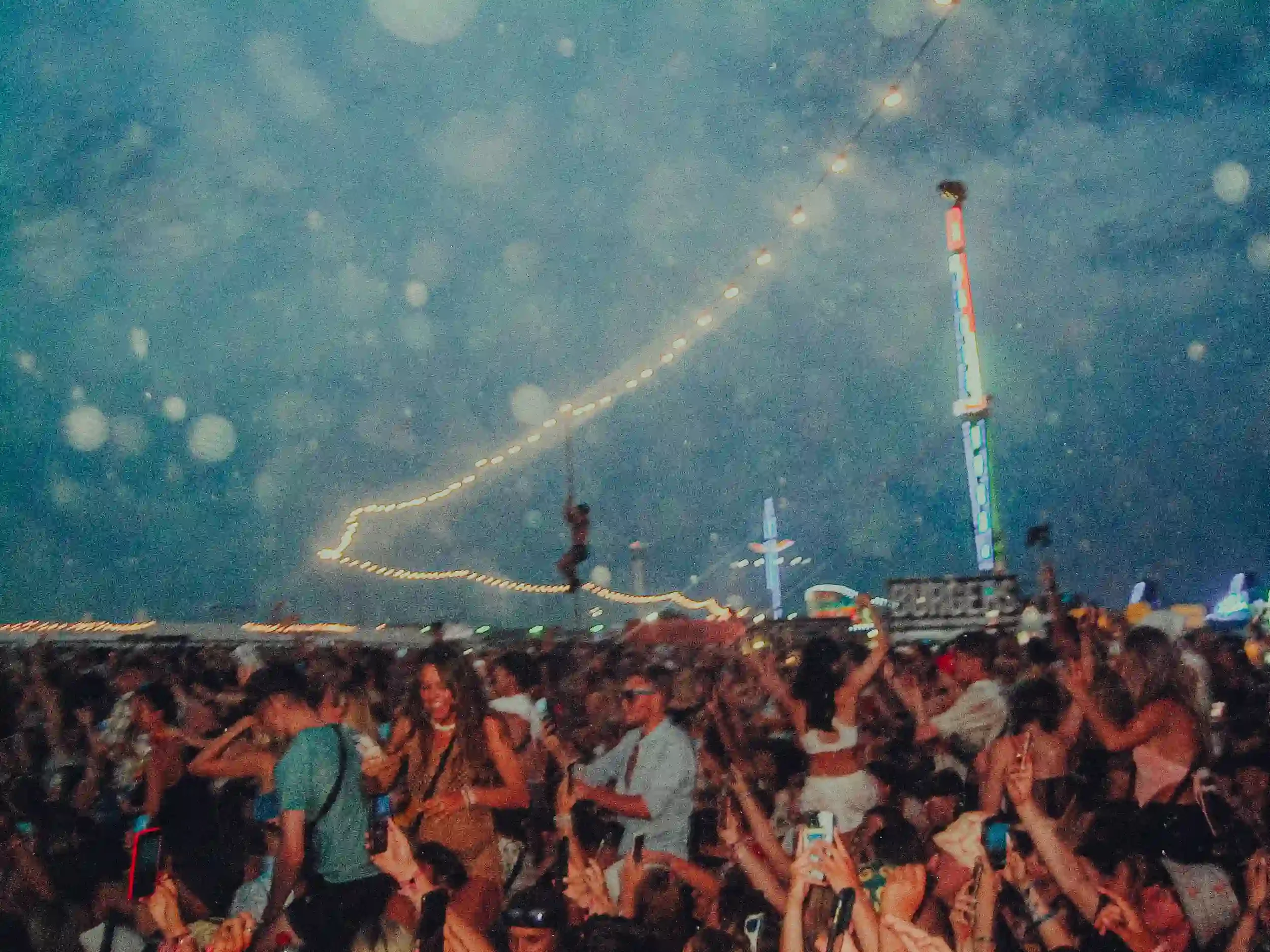 A grainy, atmospheric night shot of a massive crowd at Boardmasters Festival in Newquay. Countless people are cheering with their hands in the air under a string of glowing warm lights, while a tall, blue-lit fairground ride towers in the background 