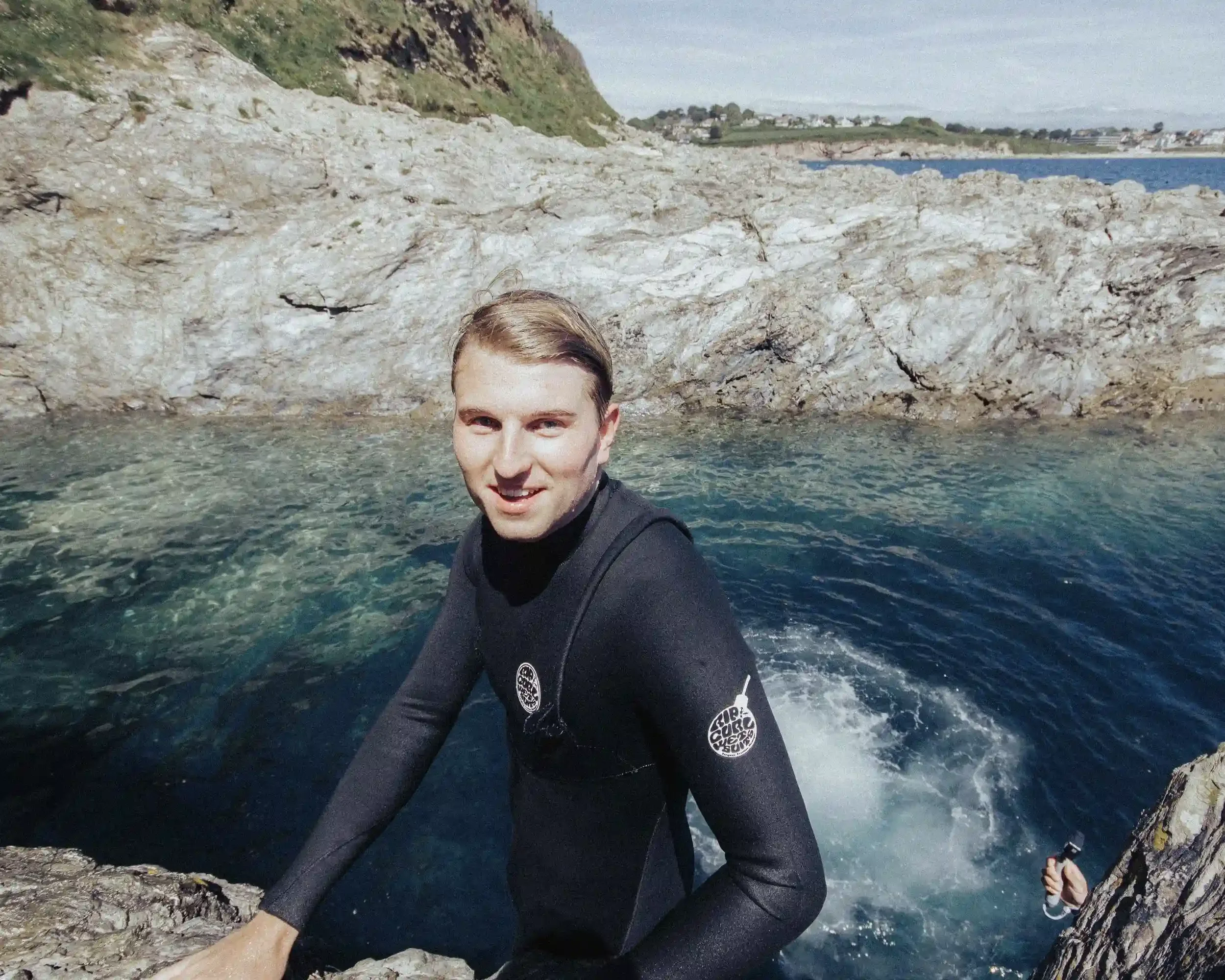 A sharp, close-up portrait of a man smiling in a black Rip Curl wetsuit, identified as the 'Ice Man' on the 'All Things Silly' music video set. He is standing against a backdrop of clear blue water and jagged grey cliffs, with a splash of water visib