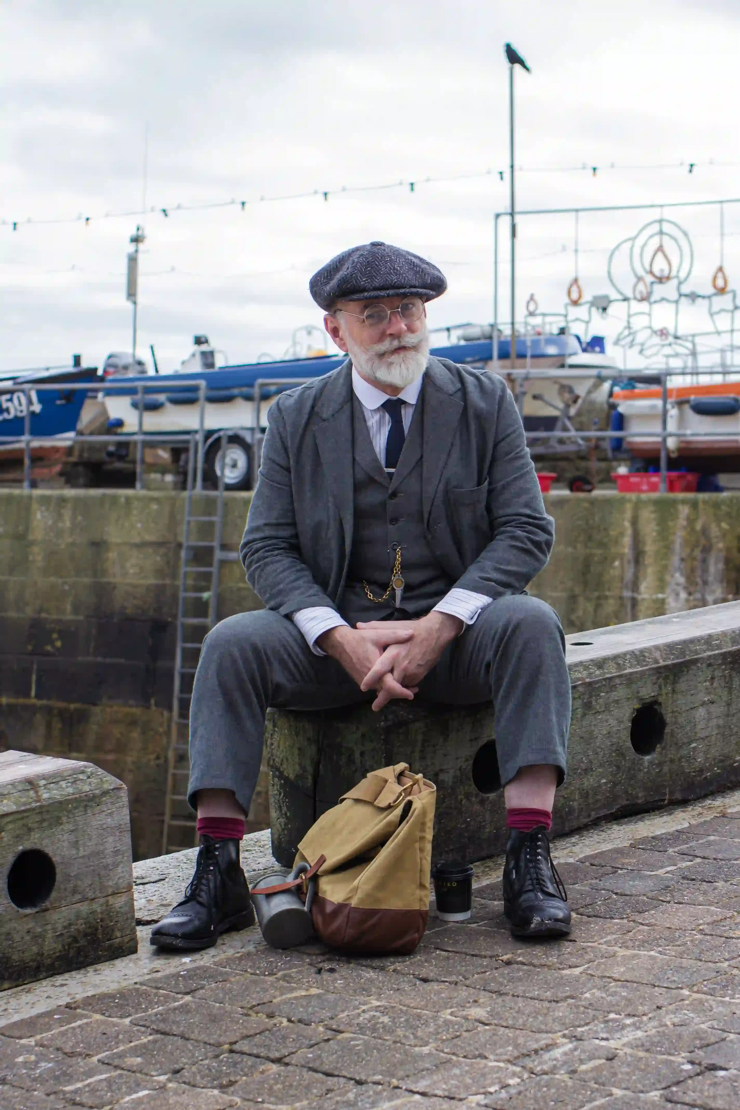 An older man with a neatly groomed white beard, round glasses, and a grey herringbone flat cap sits on a weathered wooden beam at St Ives harbor. He is wearing a formal grey three-piece suit, a dark tie, and burgundy socks with black boots, with a ta
