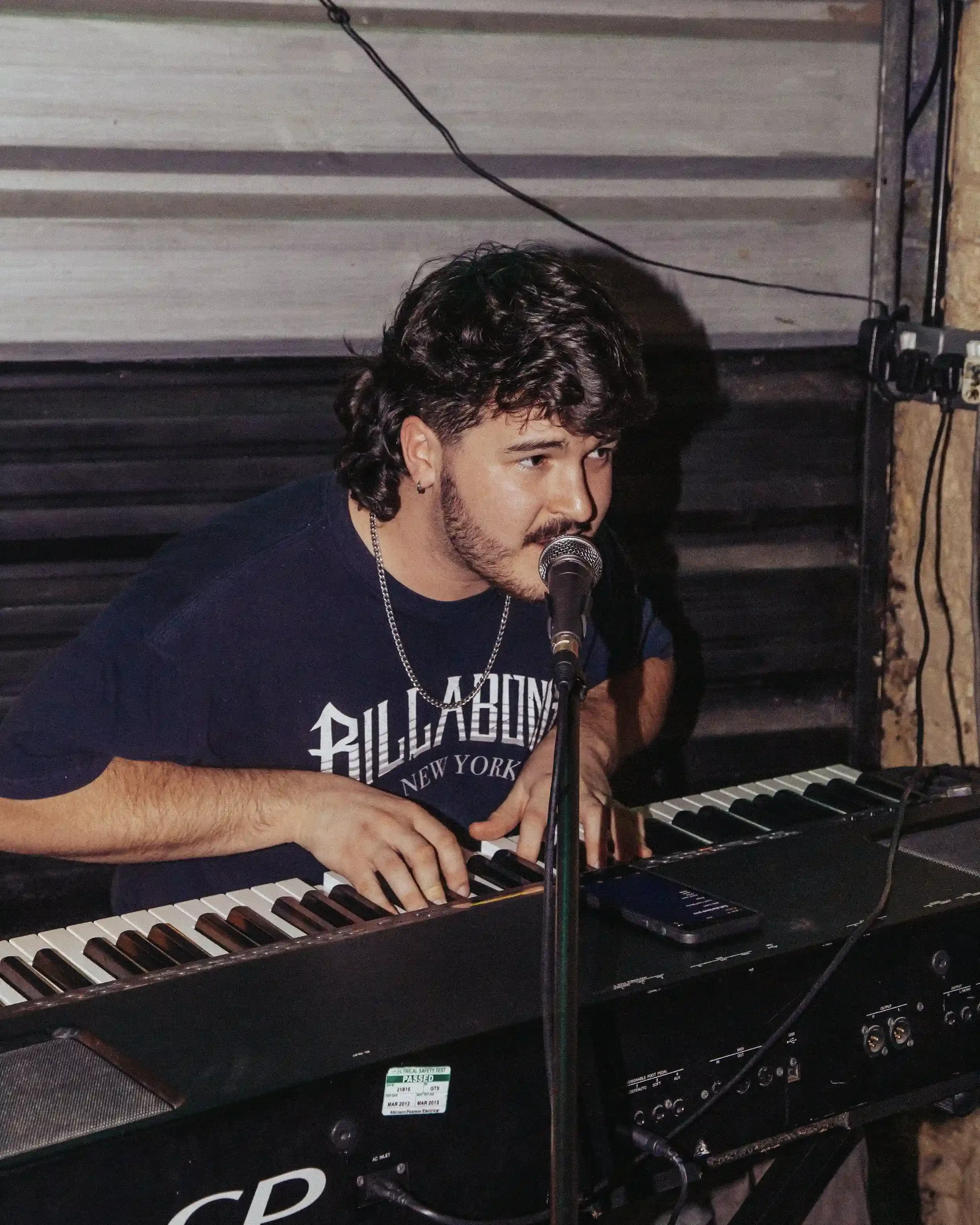 A high-contrast, candid photograph of a man with a mustache and wavy hair playing a black digital keyboard and singing into a microphone. He is wearing a dark Billabong New York t-shirt and a silver chain. The background is a dark, industrial metal w