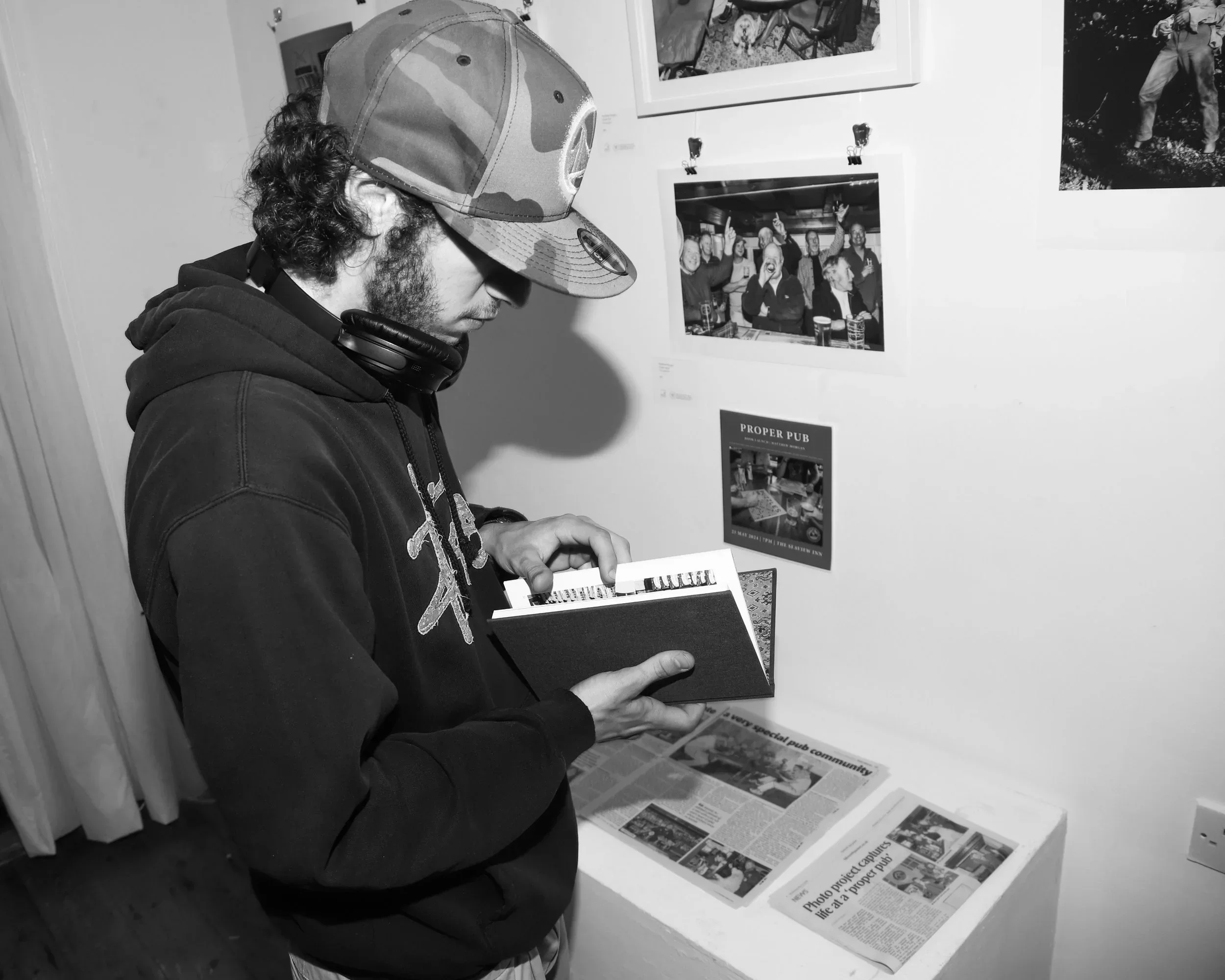 A black-and-white candid photograph of a young man with a beard, wearing a camo baseball cap and headphones, flipping through a small, dark-covered book titled 'Proper Pub.' He is standing at a white gallery plinth that displays newspaper clippings a