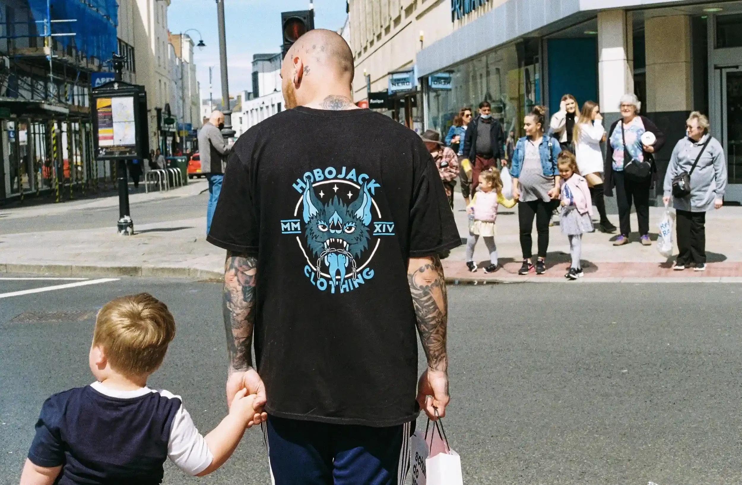 A busy street scene in Cheltenham during the 2021 lockdown recovery, showing people walking and socializing in a public square, by Matthew Morgan for the series 'Restricted Travel'.