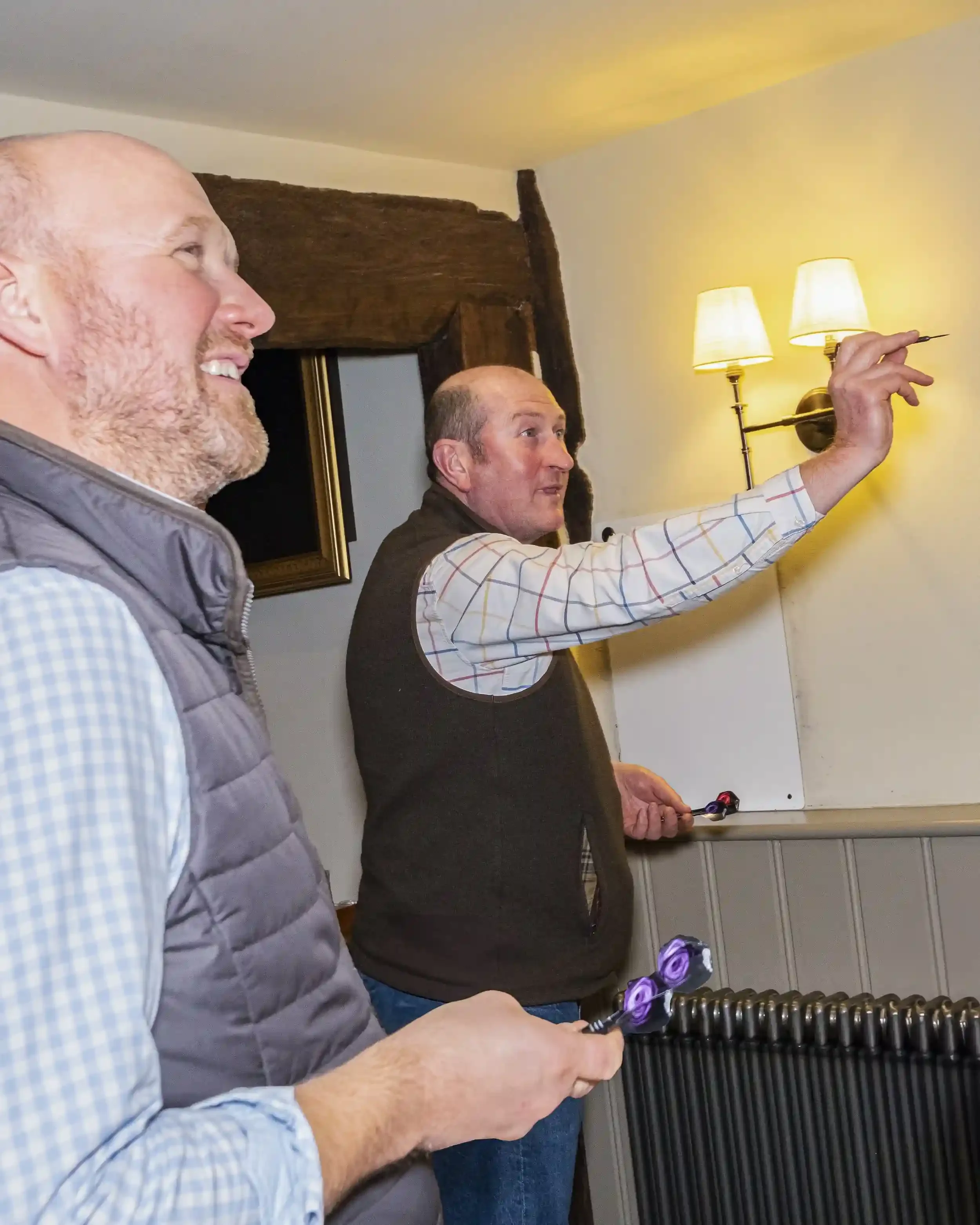 A candid interior photograph of two men playing darts at The Crown Inn, Lea. In the foreground, a man in a blue gingham shirt and grey quilted gilet smiles as he watches the game. In the background, another man in a checked shirt and brown fleece ves