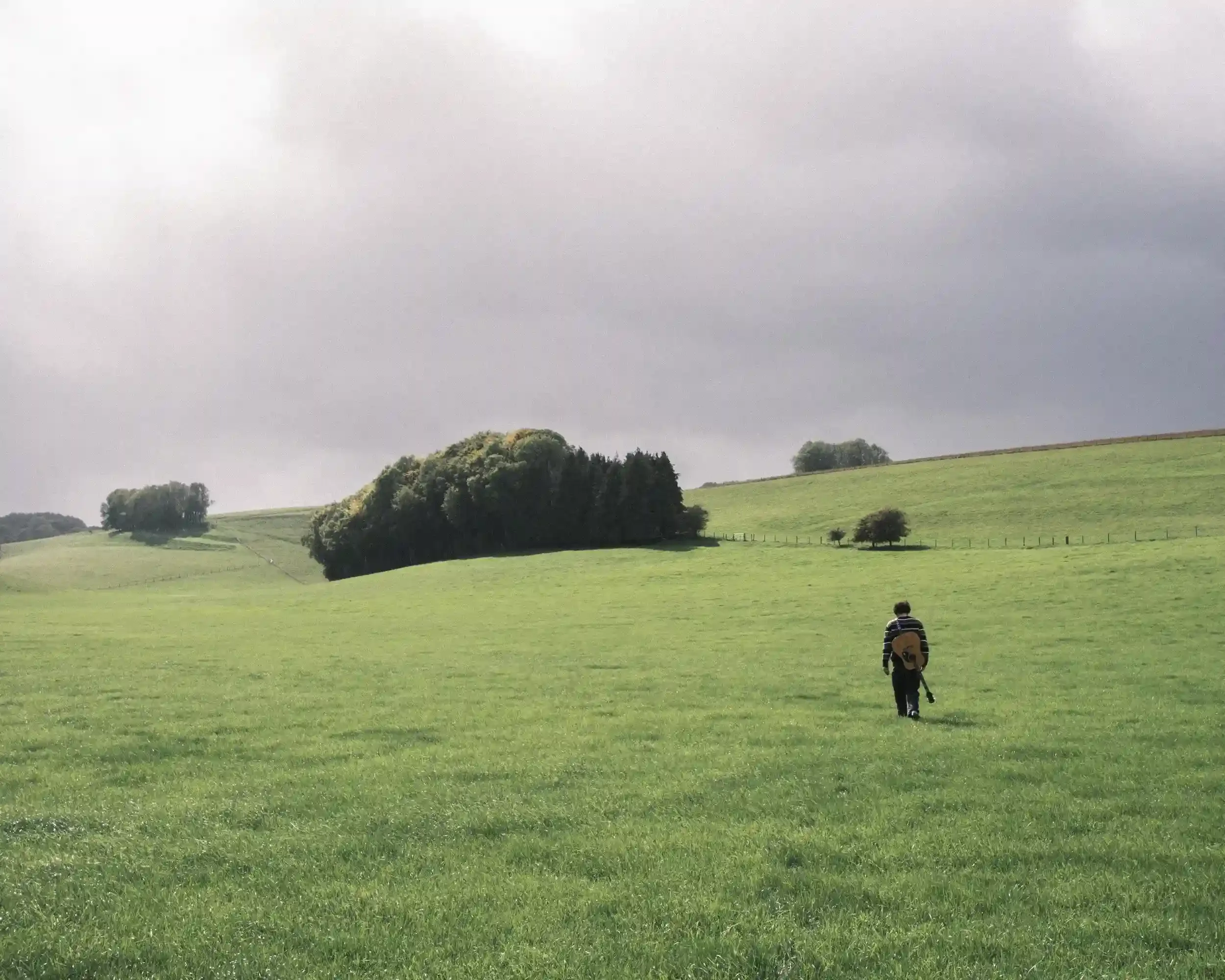 A cinematic landscape photograph of singer-songwriter Ned Holland walking away from the camera into the center of a vast, rolling green field in Salisbury. Holland, carrying an acoustic guitar over his shoulder, is a small figure against the immense 