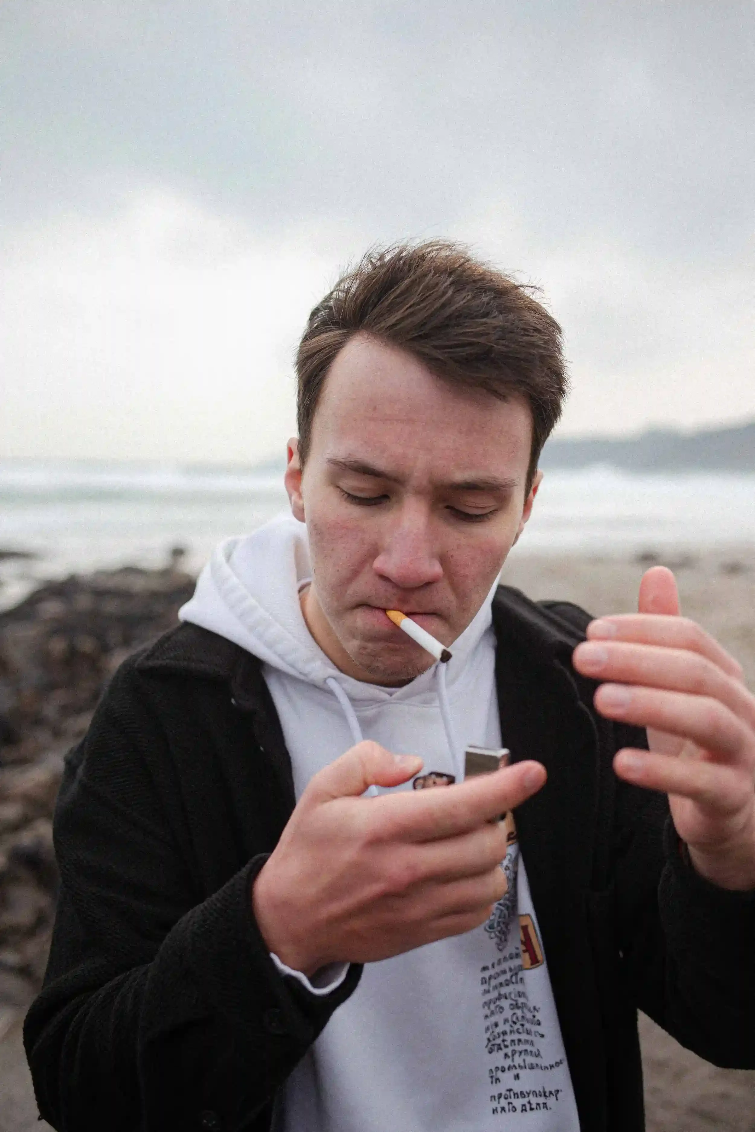 A medium close-up colour unit still of a young man with brown hair on a blurred beach. He is wearing a black open shirt over a white hoodie and is looking down, focused on using a silver lighter to light a cigarette in his mouth. His hands are slight