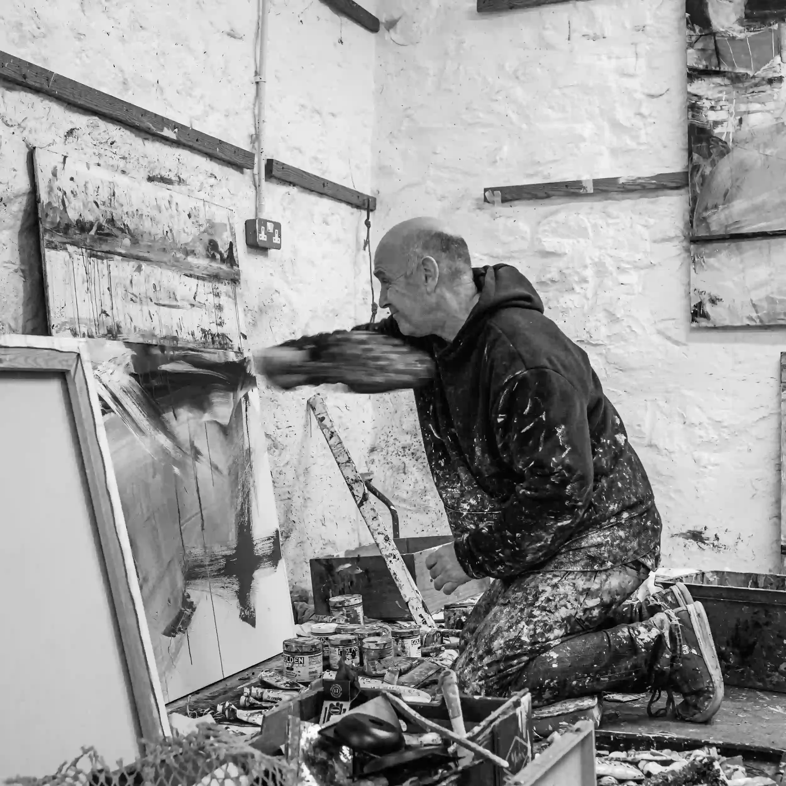 A black-and-white action photograph of artist David Mankin kneeling on his studio floor in Cornwall. His arm is a blurred arc of motion as he sweeps a tool across a canvas to create a large, dark abstract stroke. He is wearing paint-splattered clothi