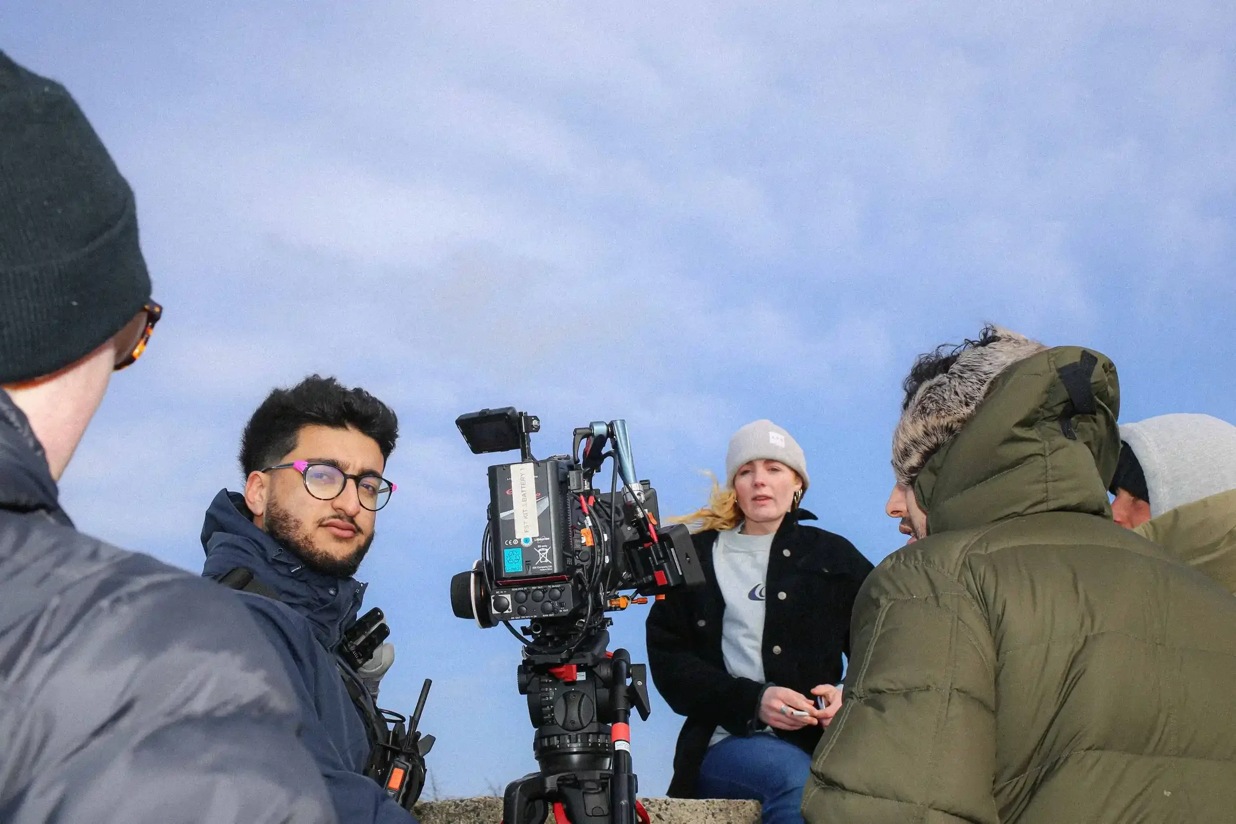 A behind-the-scenes colour photograph shot from a low angle on the set of 'Lighter.' A female actor wearing a grey beanie is seated directly behind a large cinema camera rig mounted on a tripod. Two crew members in heavy jackets are seen in the foreg