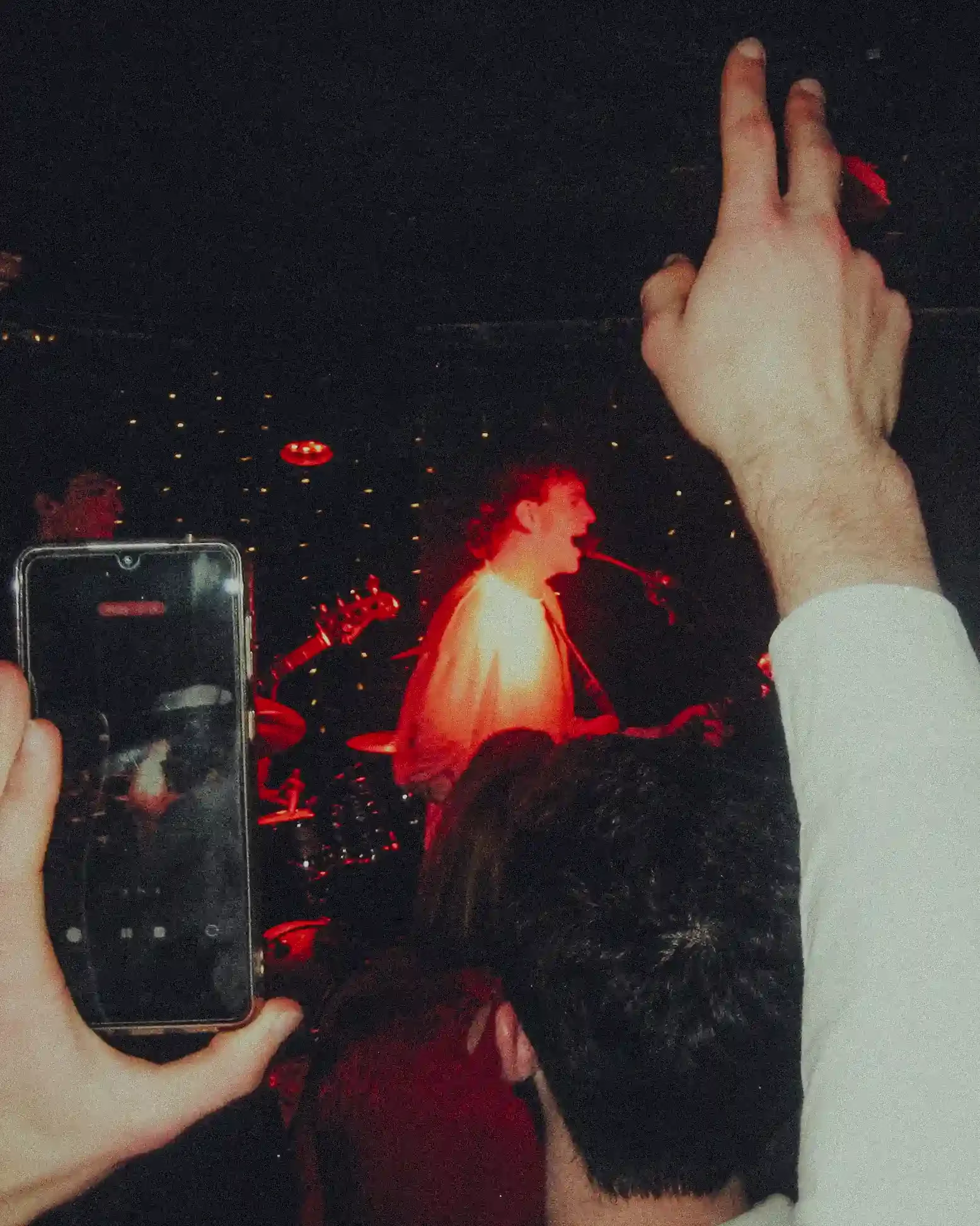 A high-grain, atmospheric photograph taken from the perspective of a crowd member at The Troubadour, London. In the left foreground, a hand holds up a smartphone with a lit screen, recording the performance. In the center, Tom Scamell is seen in prof