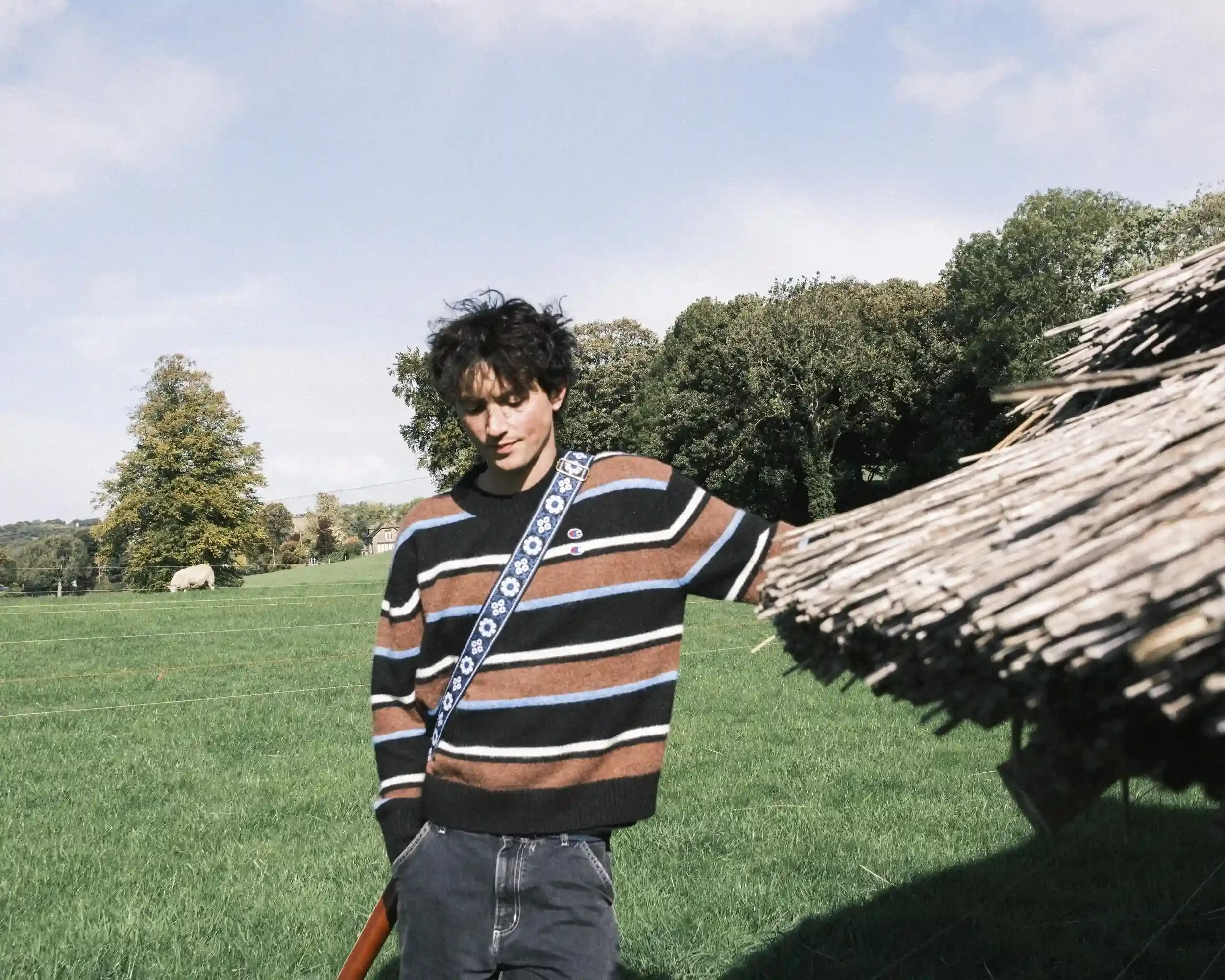 A bright, outdoor color portrait of musician Ned Holland for his single 'Bobbys.' Holland is leaning against the edge of a rustic thatched roof, looking down with a soft smile. He wears a horizontal-striped jumper in black, brown, and light blue, wit