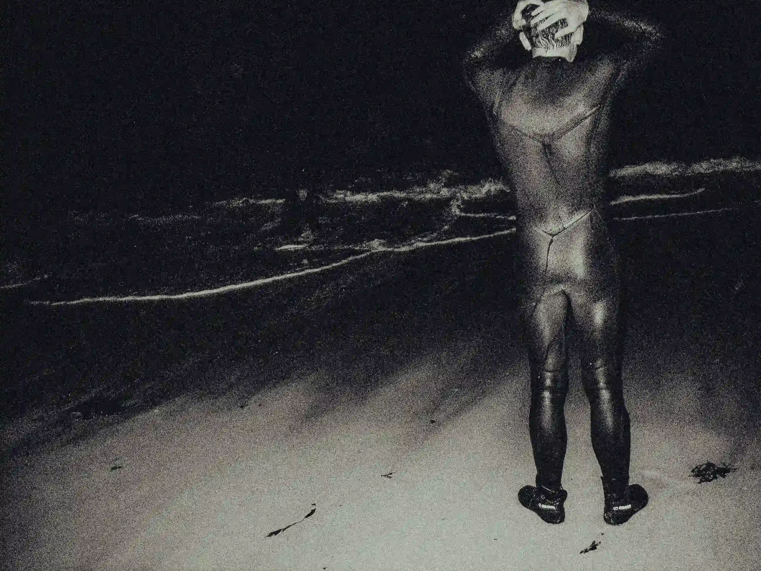 A grainy, high-contrast black-and-white photograph of a person from behind, wearing a full black wetsuit and booties. They are standing on a dark beach with their hands on their head, looking toward a single thin line of white foam from a wave breaki