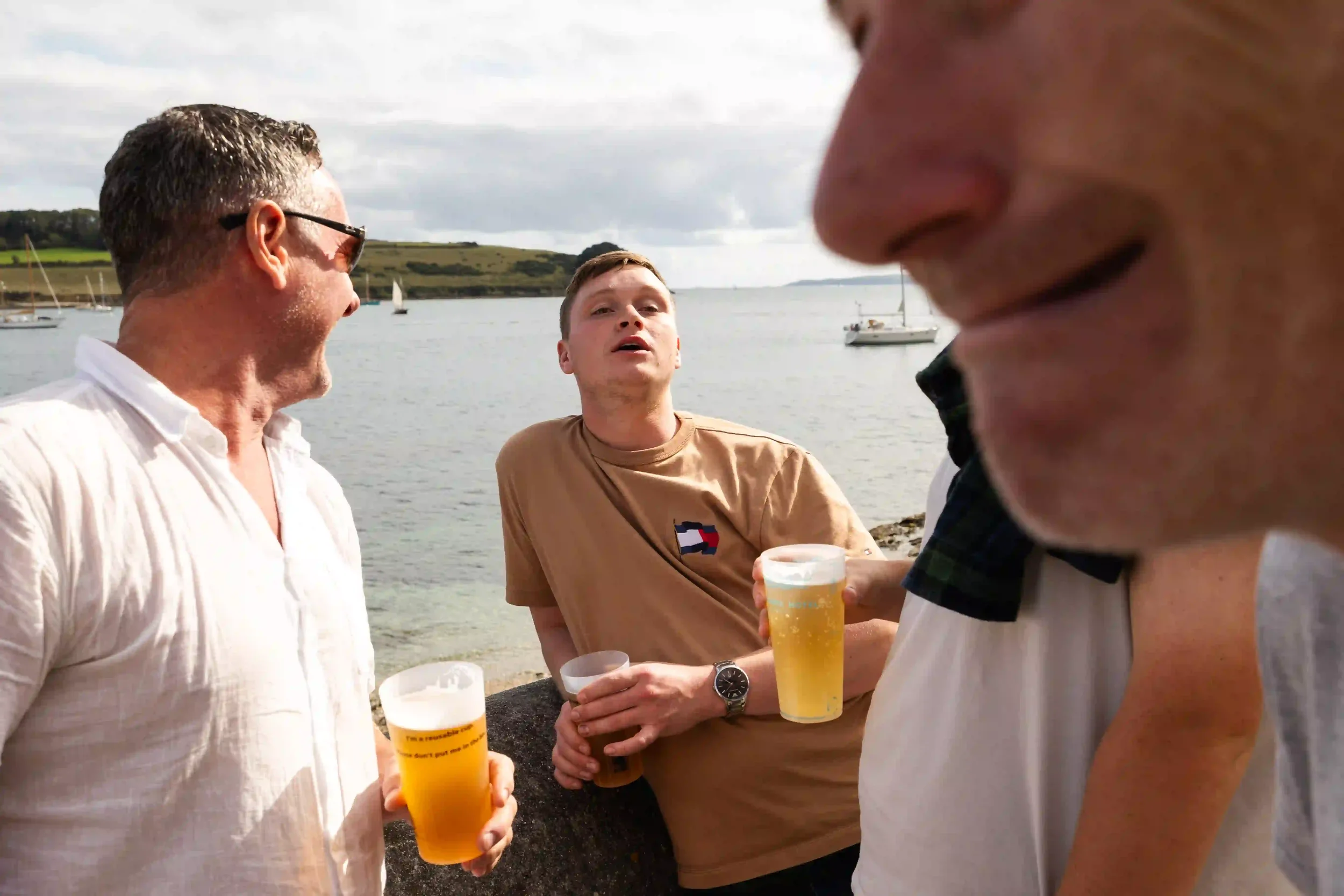 A group of men stand drinking beer in reusable plastic cups on a sunny day outside the St Mawes Hotel. In the background, white sailing boats sit on the calm blue water of the Falmouth estuary under a cloudy sky, with a close-up, blurred face of a ma