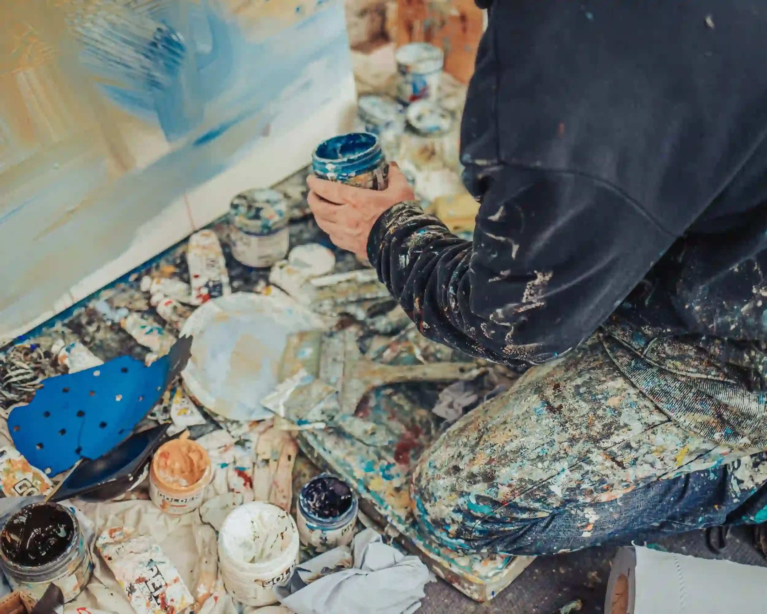 A vivid colour close-up of artist David Mankin's hands and legs as he works on the floor of his Cornwall studio. He holds a jar of bright blue paint, surrounded by an array of open acrylic paint containers, used brushes, and blue sandpaper on a paint